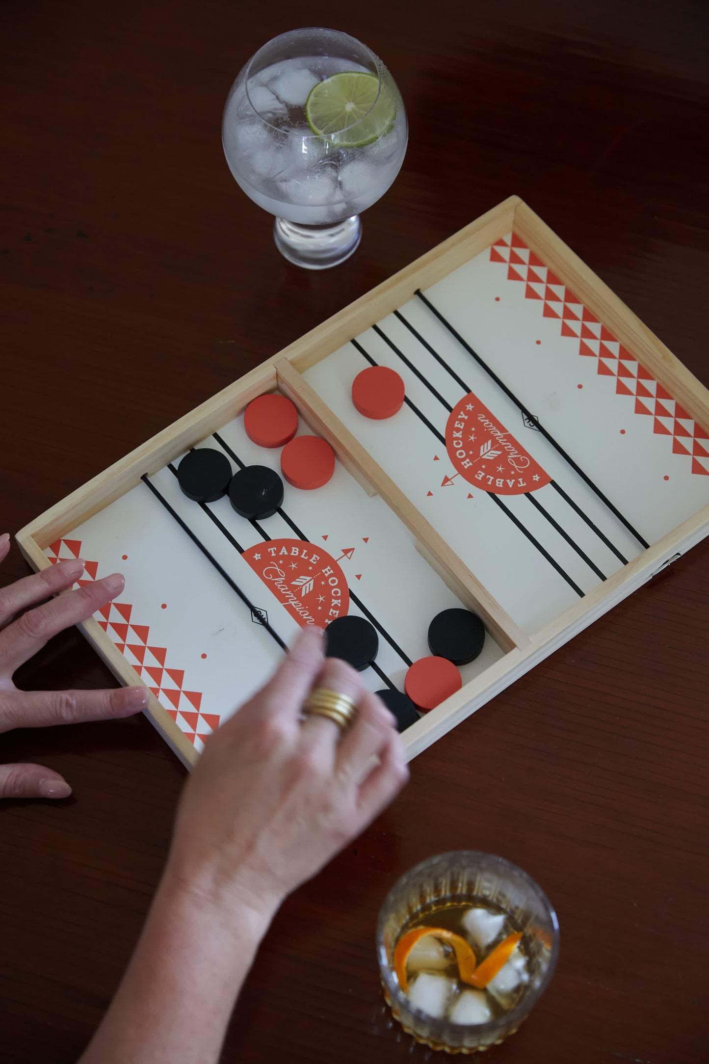 Two people play Table Hockey with black and red pieces on a wooden board. Nearby, a clear drink with lime and an amber drink with ice sit on the table, hinting at friendly competition in this elastic tabletop game.