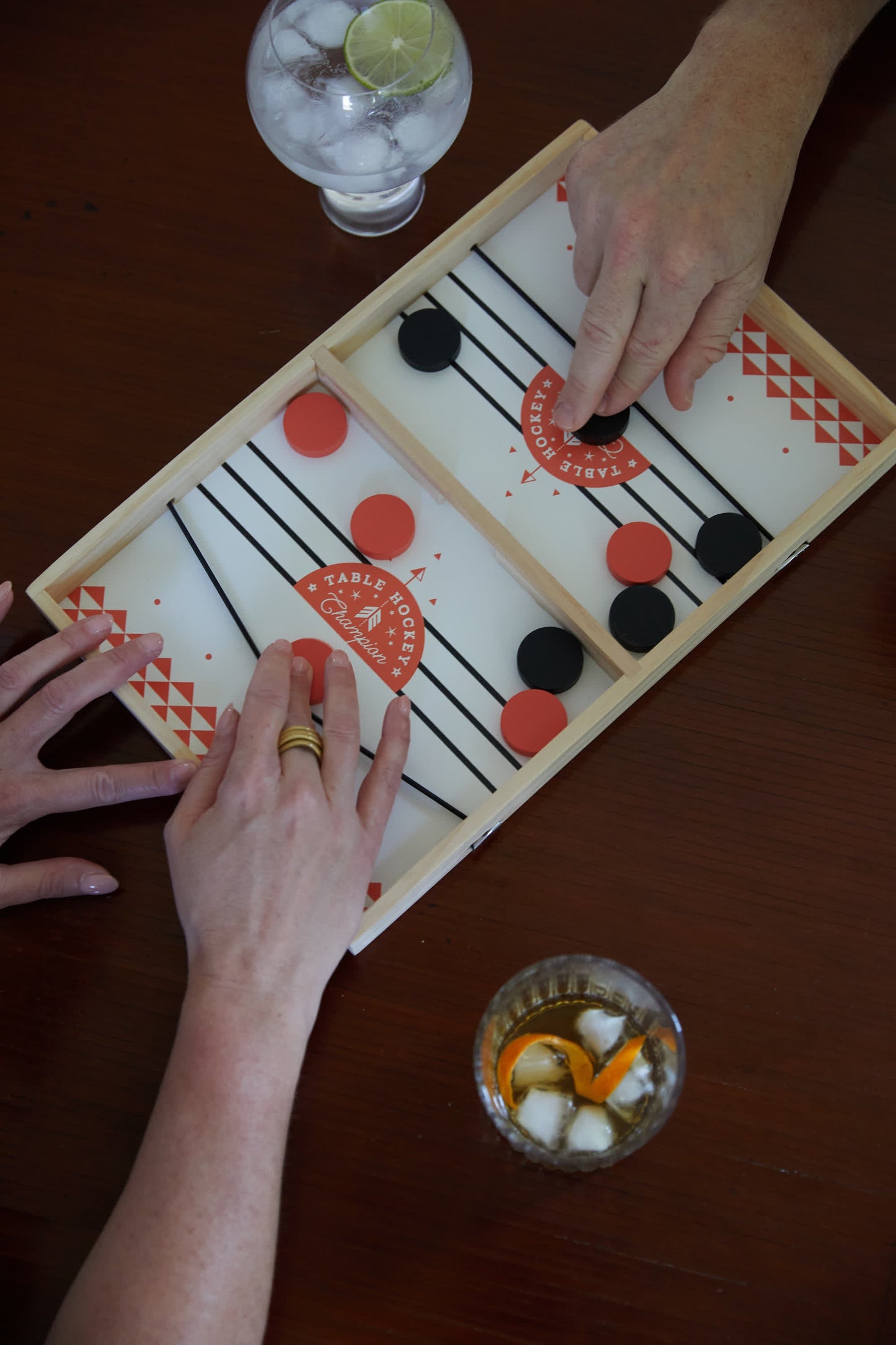 Two people play Table Hockey, a fast tabletop game with red and black discs on a wooden board using an elastic launcher. Nearby, two drinks with ice and garnish sit on the brown table.