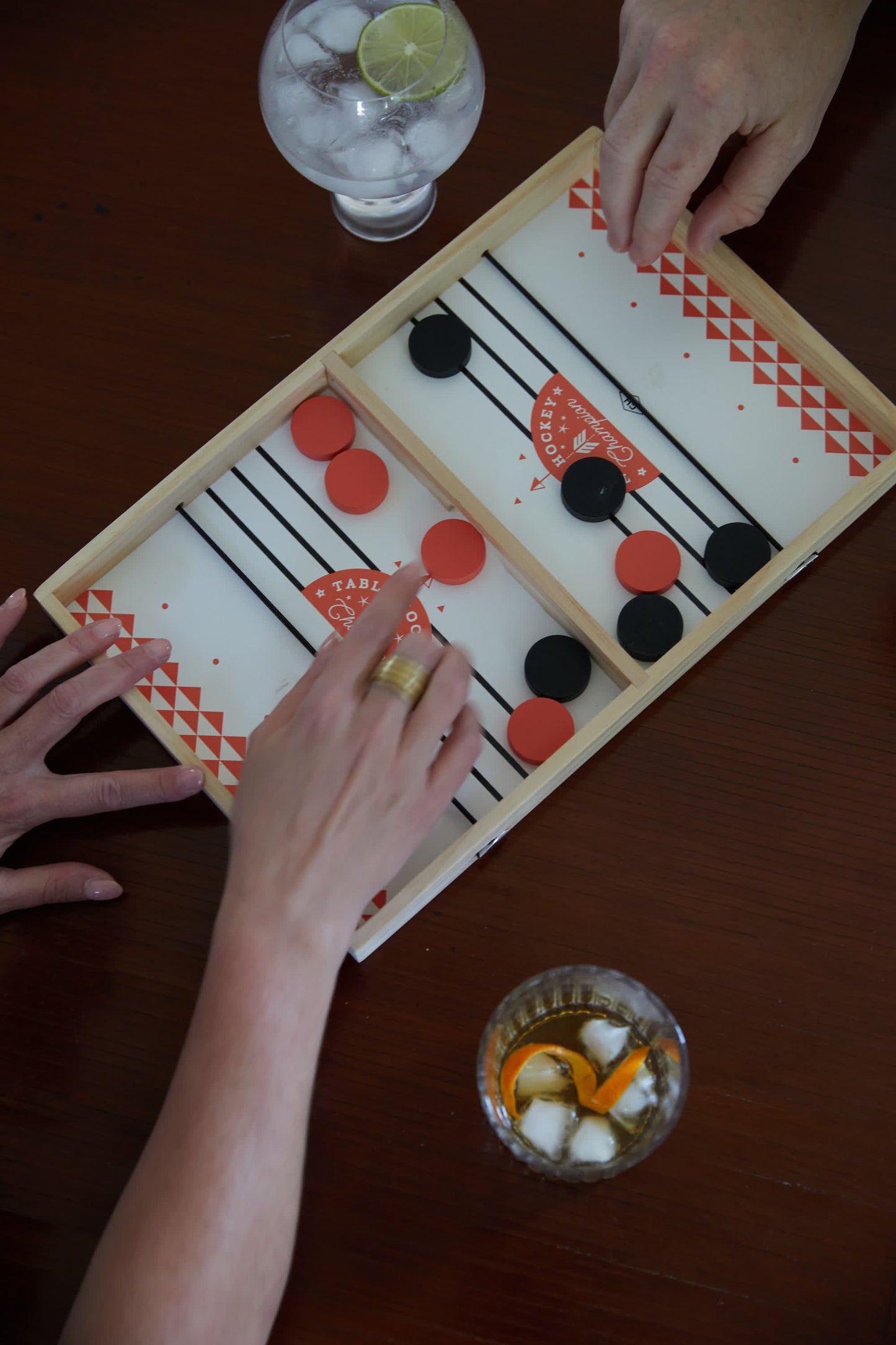 Two people play Table Hockey with black and red pieces on a wooden board. Refreshing drinks with ice and garnish rest on the brown table, adding enjoyment to their match.