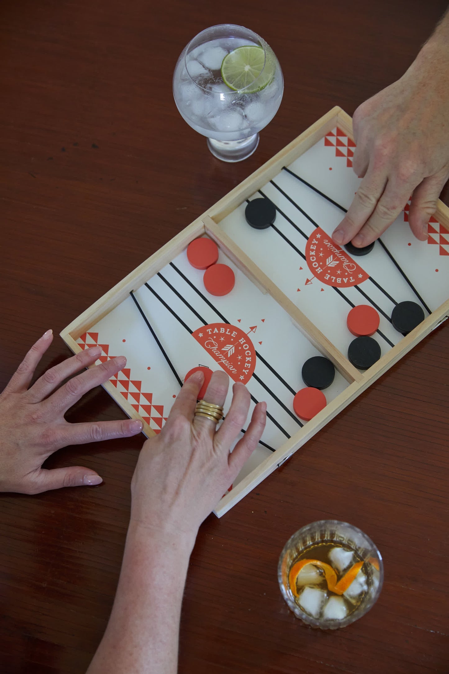 Two people play Table Hockey with black and red pieces on a wooden board. Nearby, one has a clear drink with lime and the other an amber drink with ice as they enjoy their game.