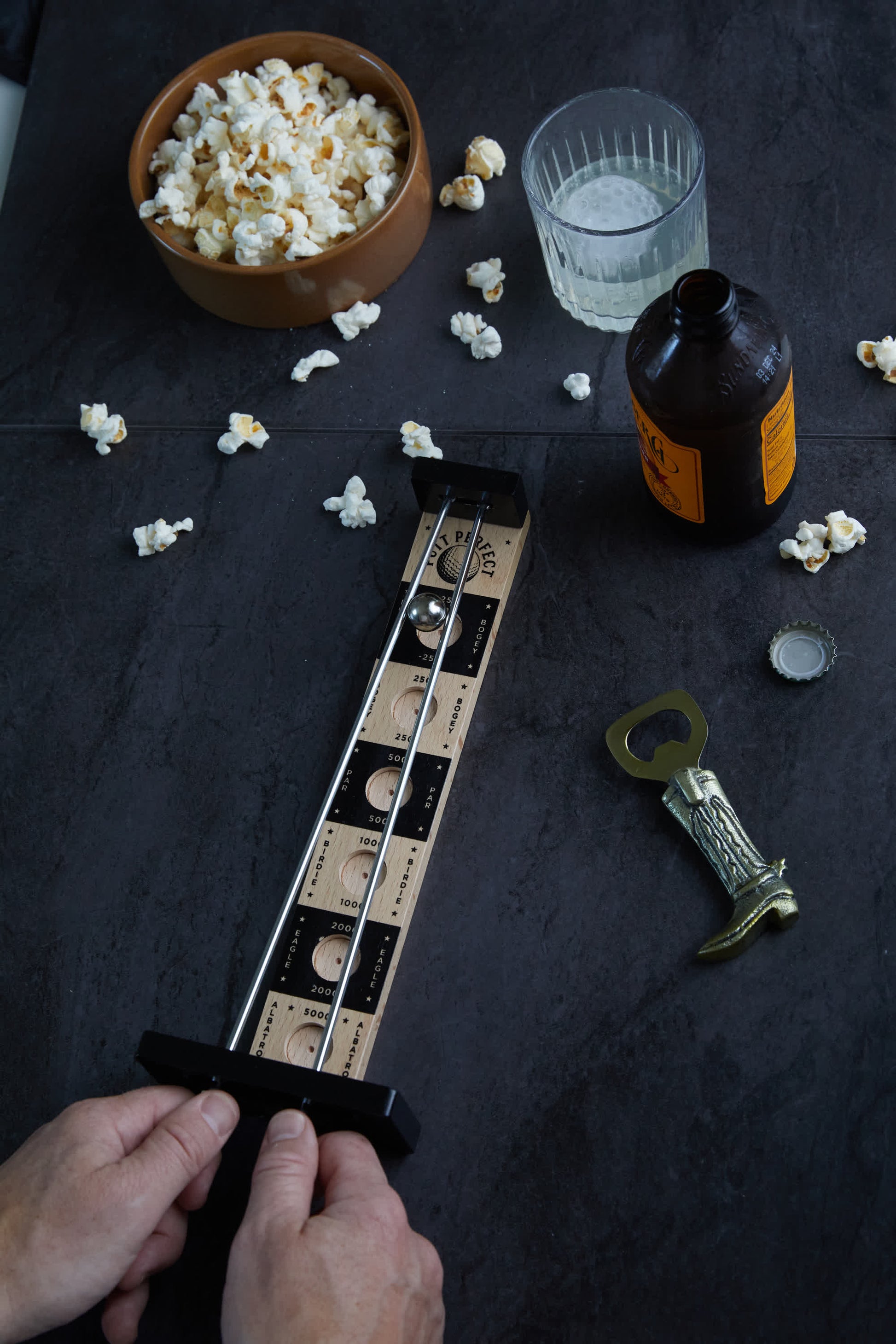 Hands holding a Putt Perfect wooden tabletop game, surrounded by scattered popcorn, a bowl of popcorn, a clear drink, a beer bottle, and a bottle opener on a dark surface.