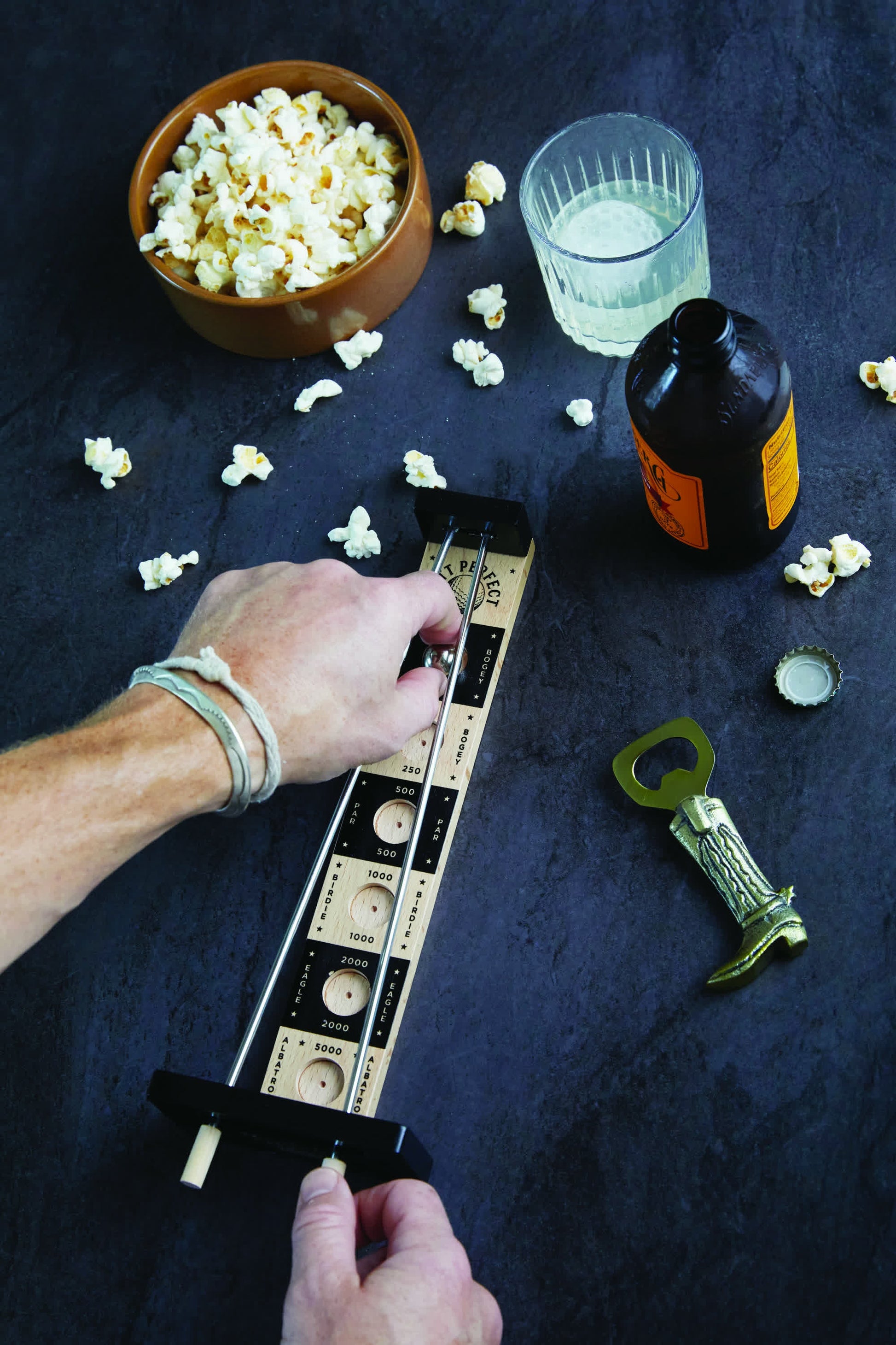 A person enjoys a tabletop pinball game on a dark surface, snacks and drinks nearby—an ideal way to unwind after sharpening their pro skills with Putt Perfect.