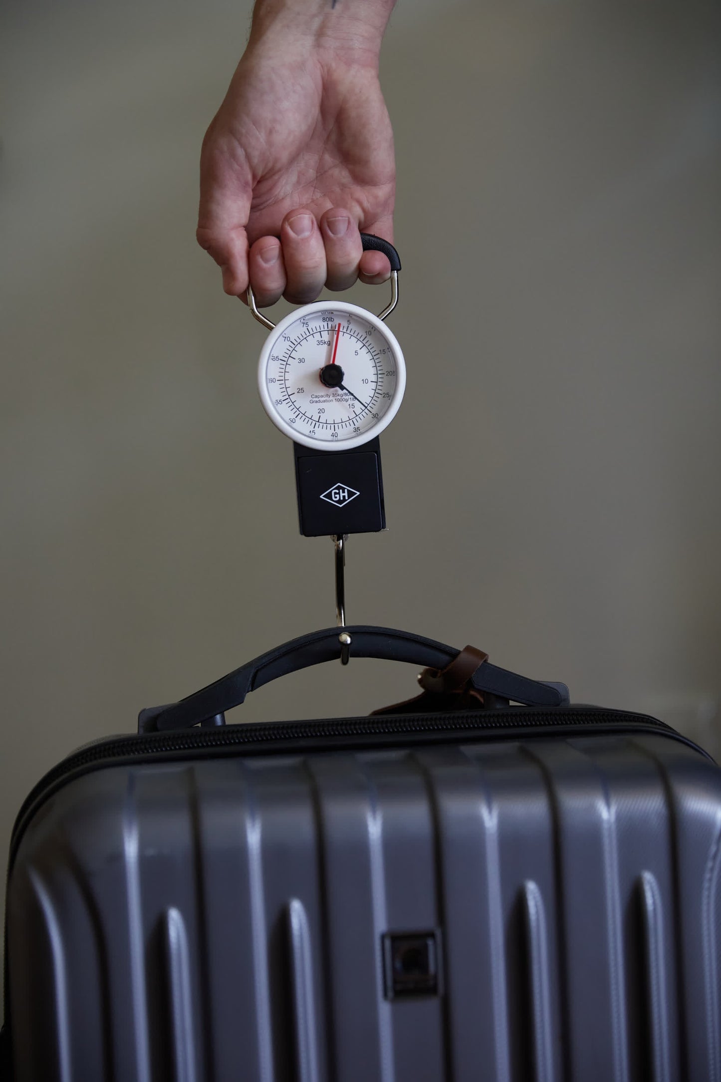 A hand holds the Travel Scale, delivering precise weight readings for a gray hard-shell suitcase—making travel packing simple and stress-free.