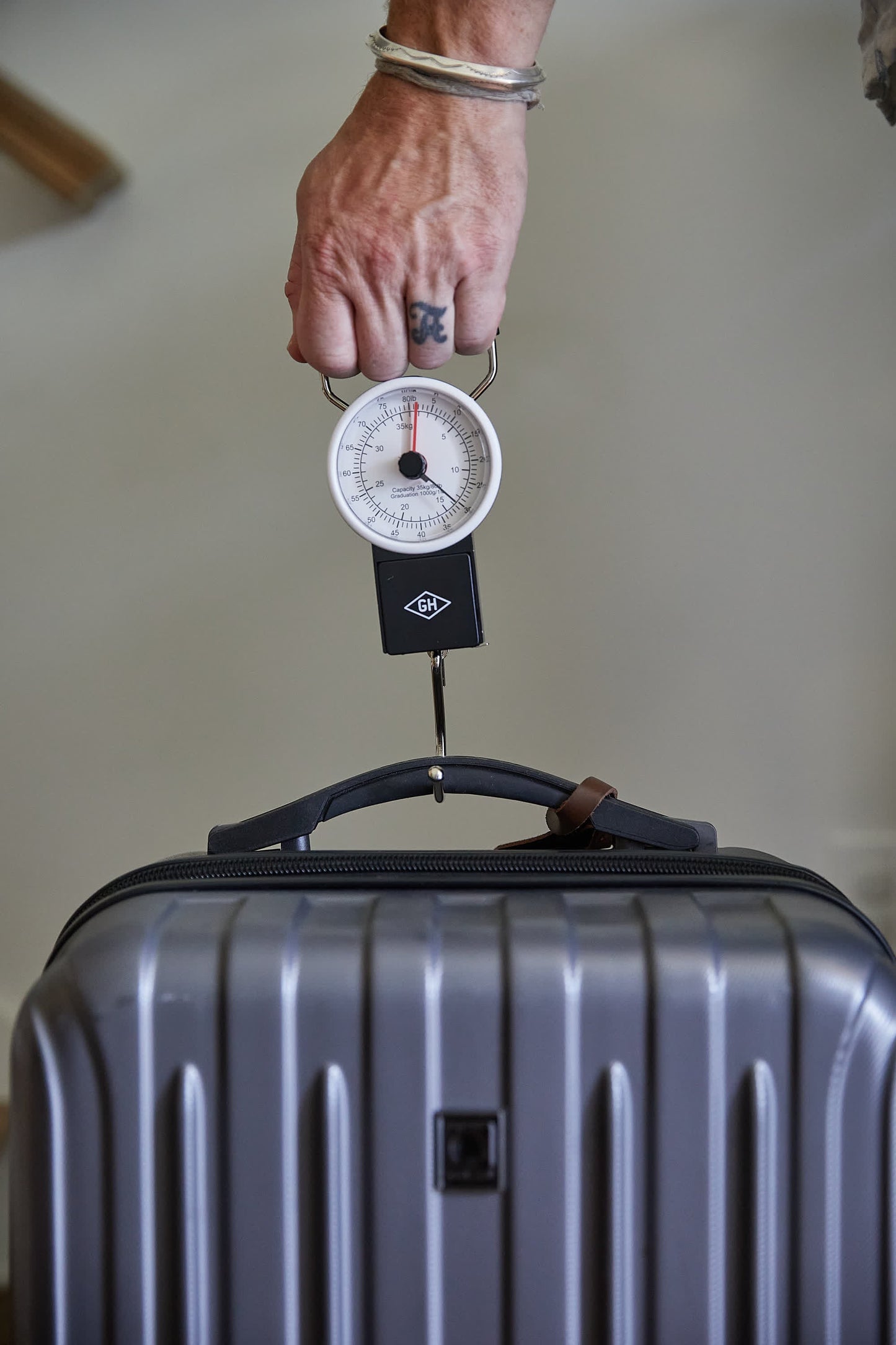 A hand holds the Travel Scale, a circular analog device that accurately measures the weight of a gray hard-shell suitcase—making it an essential travel companion.