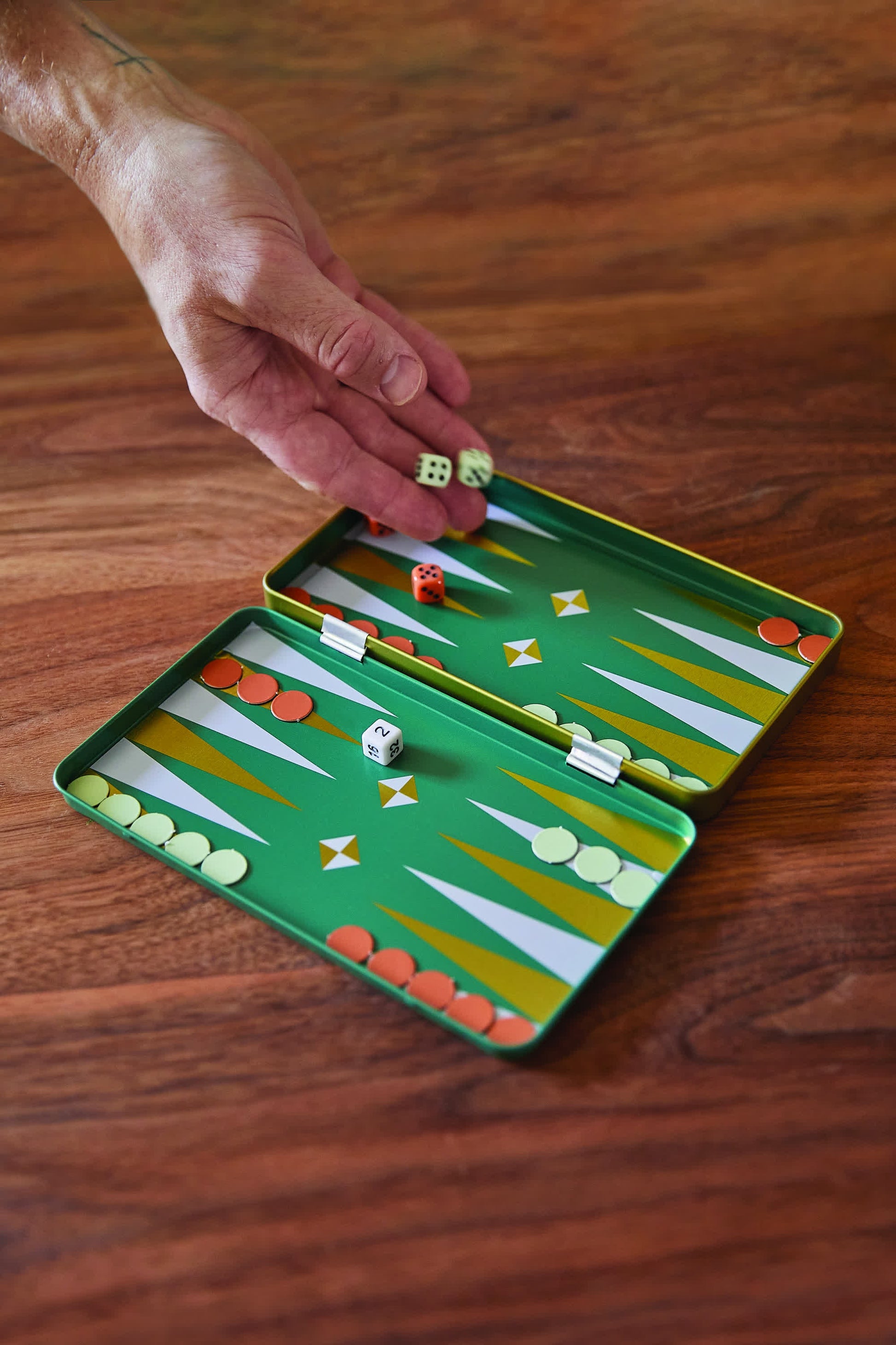 A hand rolls dice onto the Travel Backgammon set featuring arranged game pieces and a secure magnetic playing surface on a wooden table.