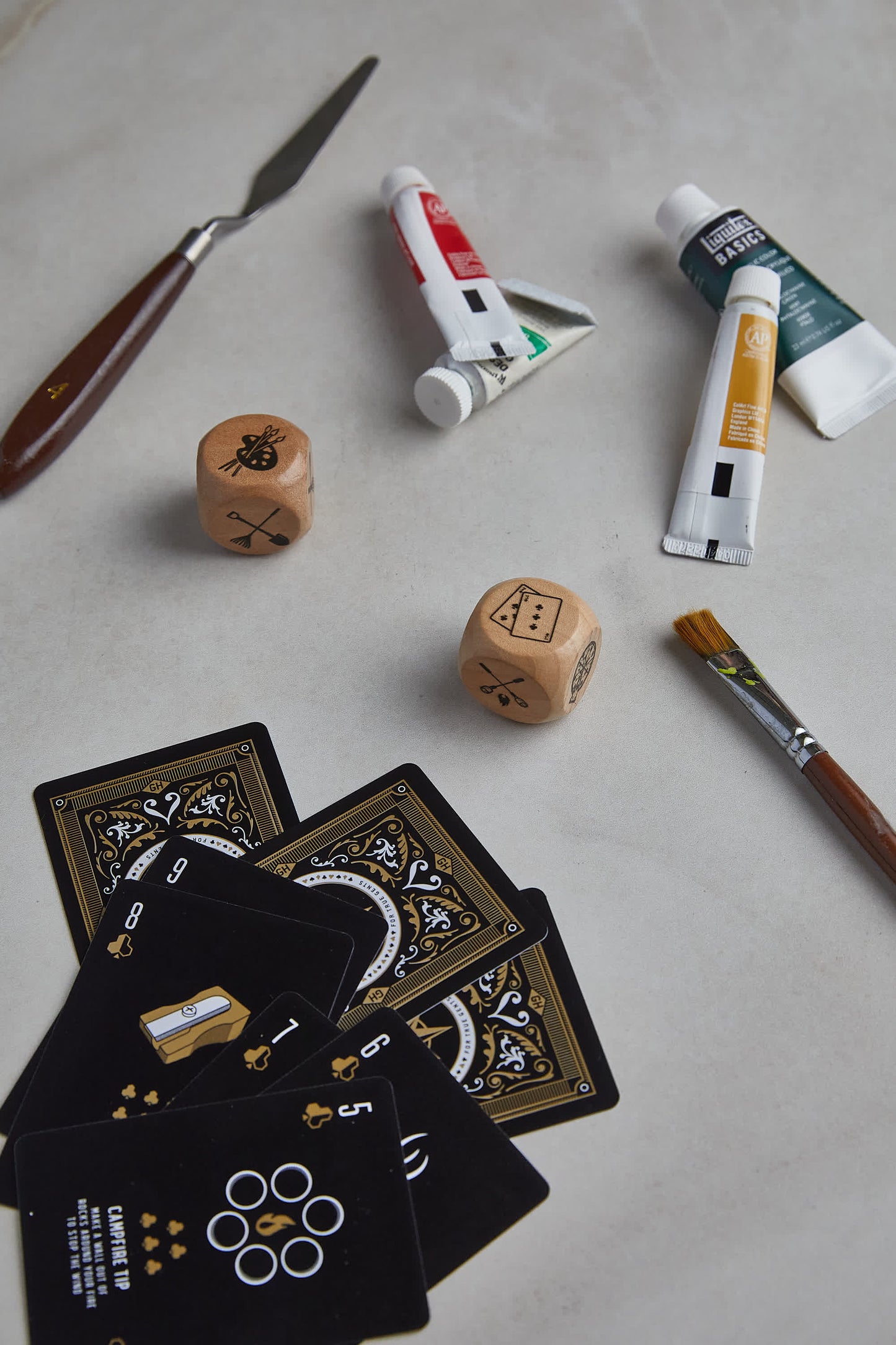 The Family Dice set, featuring two wooden dice, paint tubes, a palette knife, a paintbrush, and playing cards on a light surface, captures the essence of family fun.