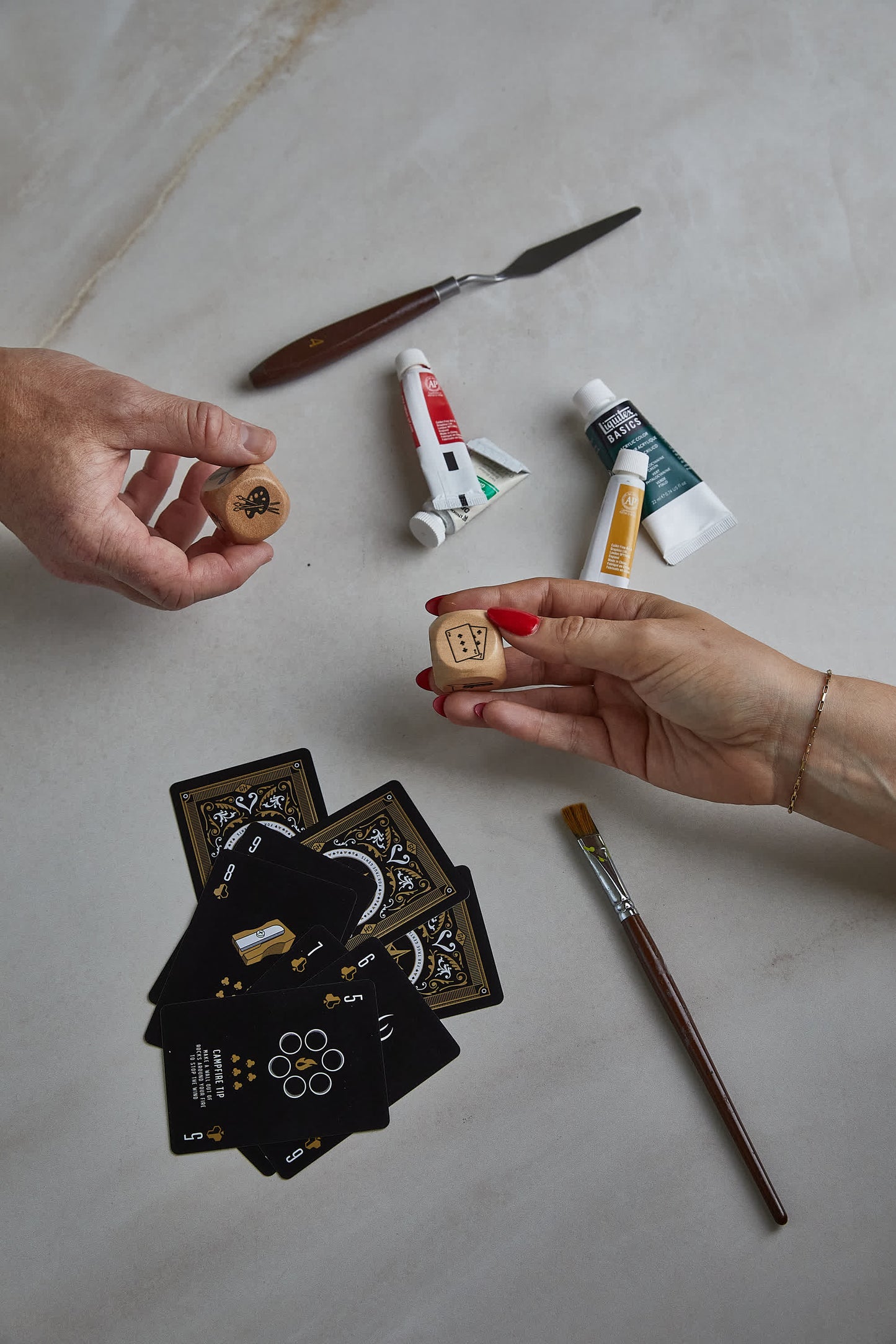 Two hands hold Family Dice over a table with playing cards, paint tubes, a palette knife, and a paintbrush—making Family Dice the perfect game for fun activities everyone in the family can enjoy together.
