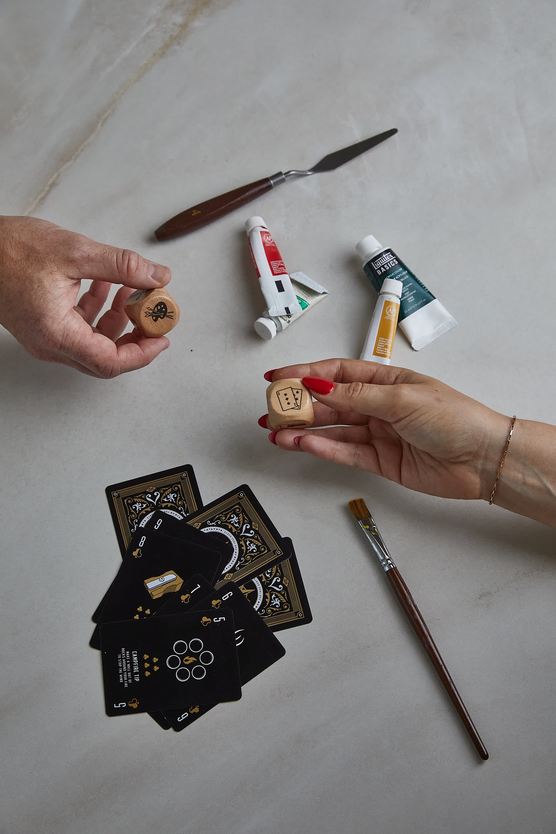 Two hands hold Family Dice over a table with playing cards, paint tubes, a palette knife, and a paintbrush—making Family Dice the perfect game for fun activities everyone in the family can enjoy together.