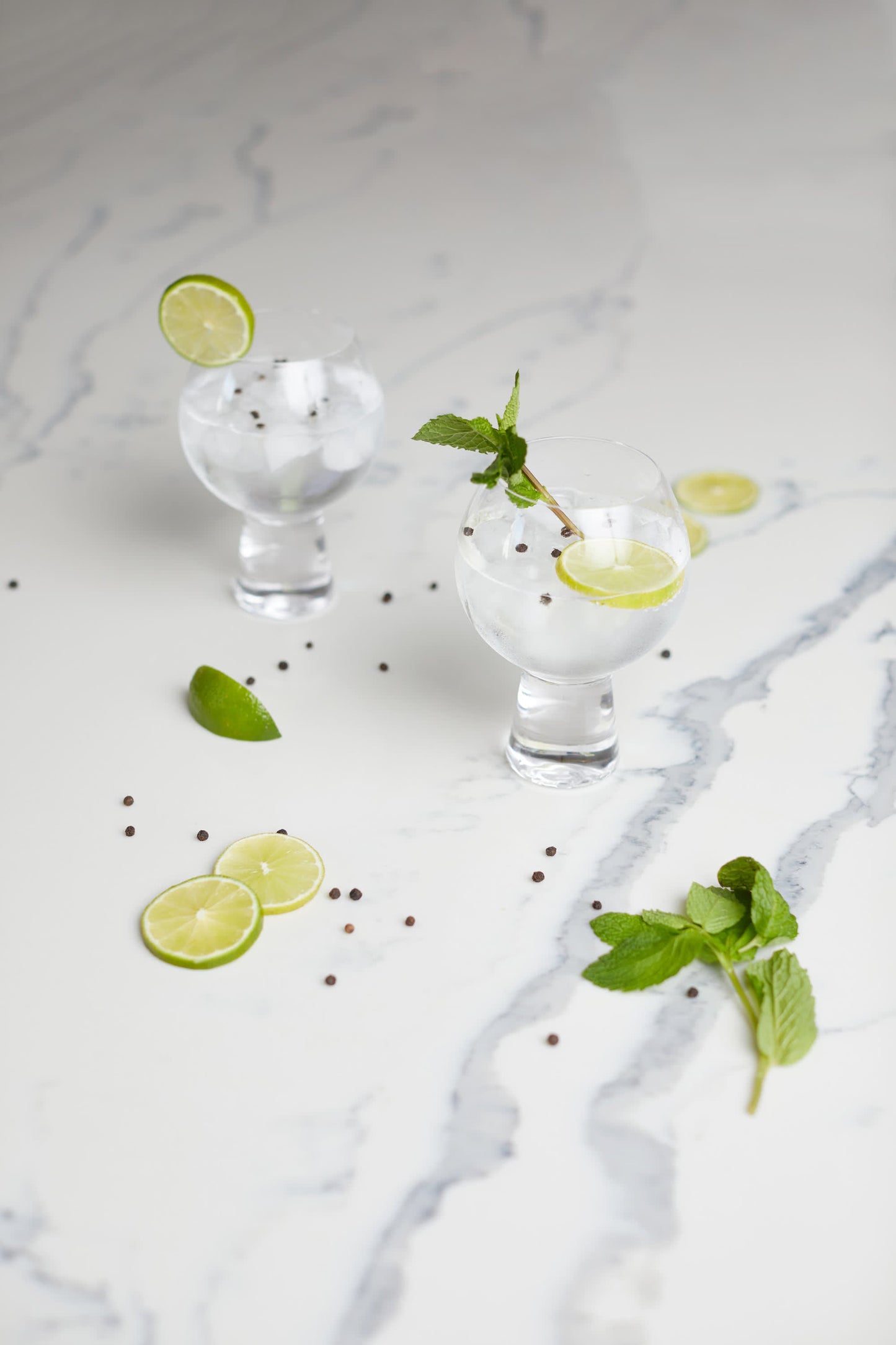 The Gin Glass - Set Of 2 features enchanting glasses filled with clear cocktails, ice, lime slices, mint sprigs, and peppercorns on marble, accented by extra lime, mint, and peppercorns for a refreshing presentation.