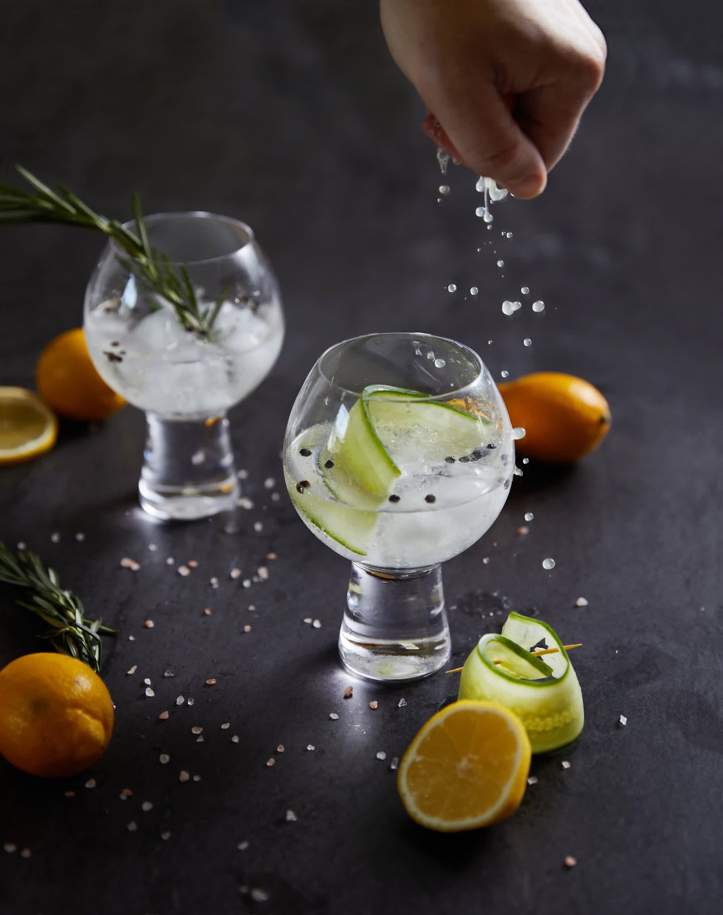 A hand sprinkles salt over two gin and tonics served in goblet-shaped Gin Glass - Set Of 2, garnished with cucumber, rosemary, and peppercorns. Citrus fruits and salt decorate the dark surface.