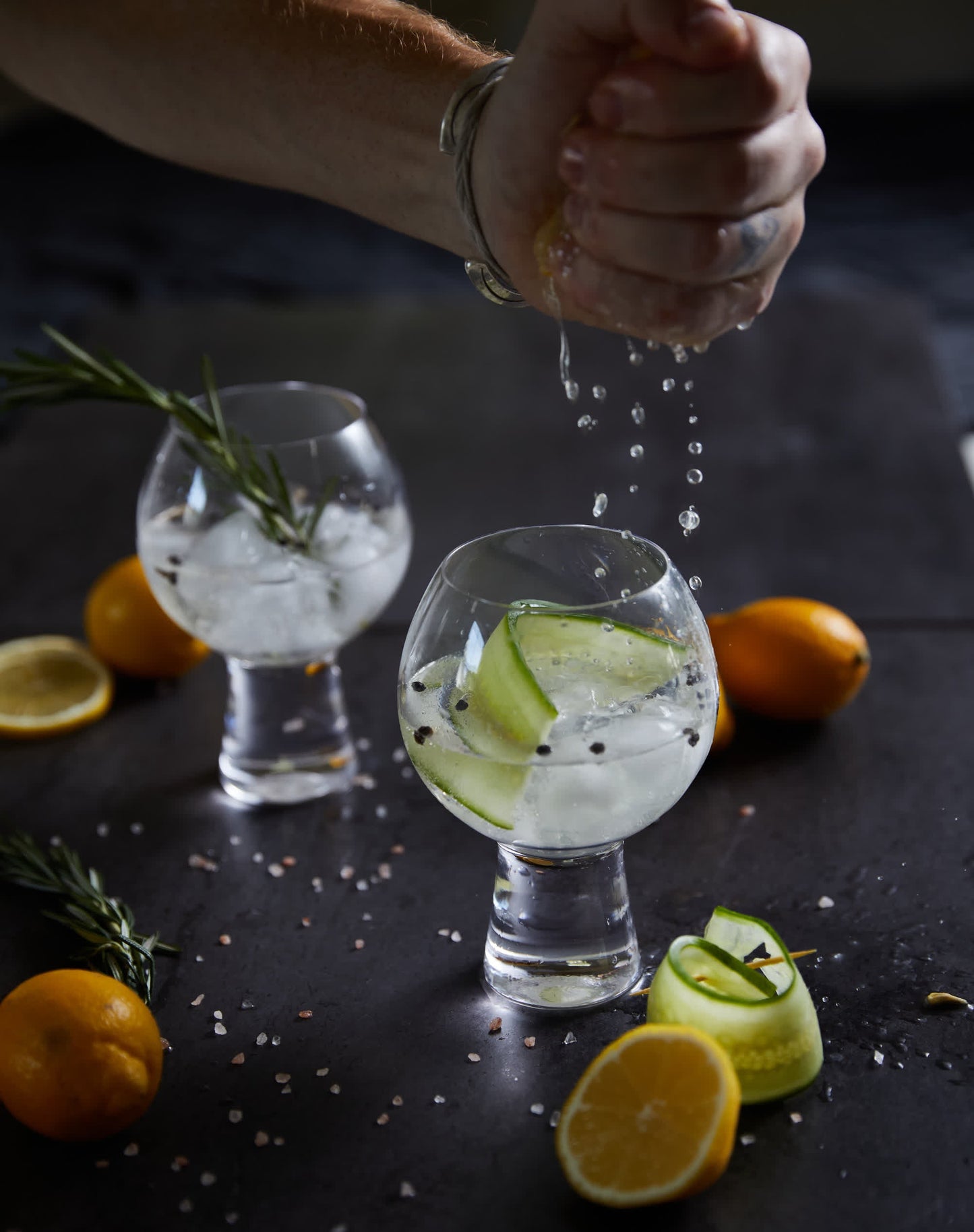 A hand squeezes lemon over two gin and tonics in enchanting Gin Glass - Set Of 2, each garnished with cucumber, rosemary, and peppercorns. The dark surface is scattered with lemons and ice.