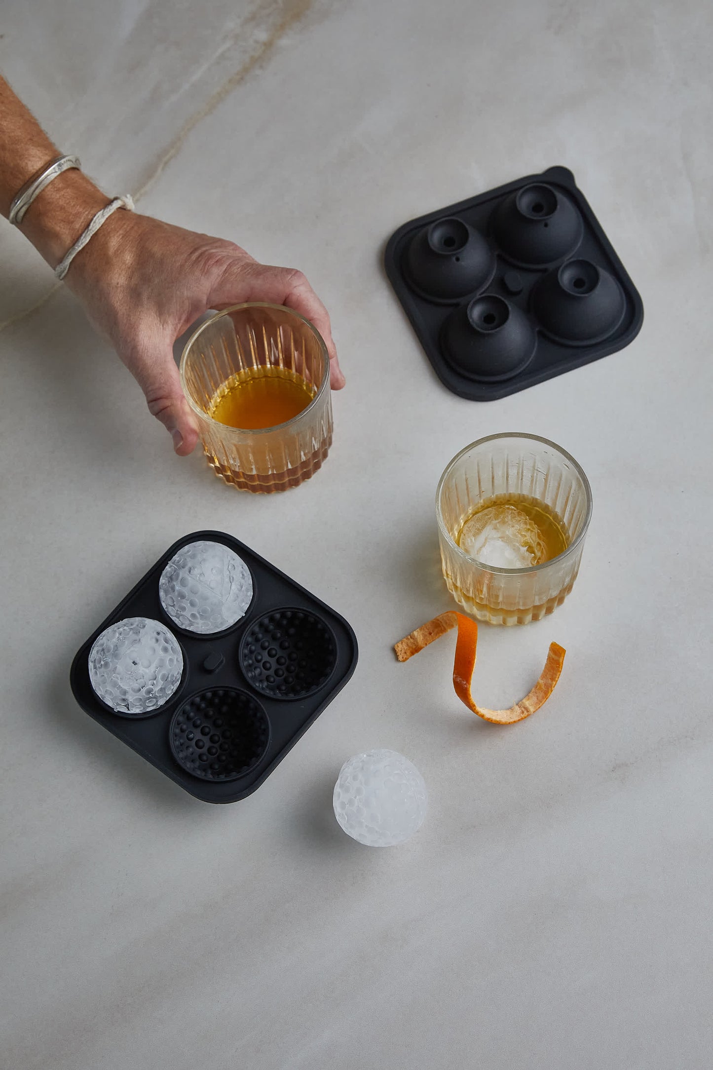 A hand holds a glass of whiskey beside another glass, an orange peel, the Golf Ball Ice Mould, three ice balls, and the mold lid on a marble surface.