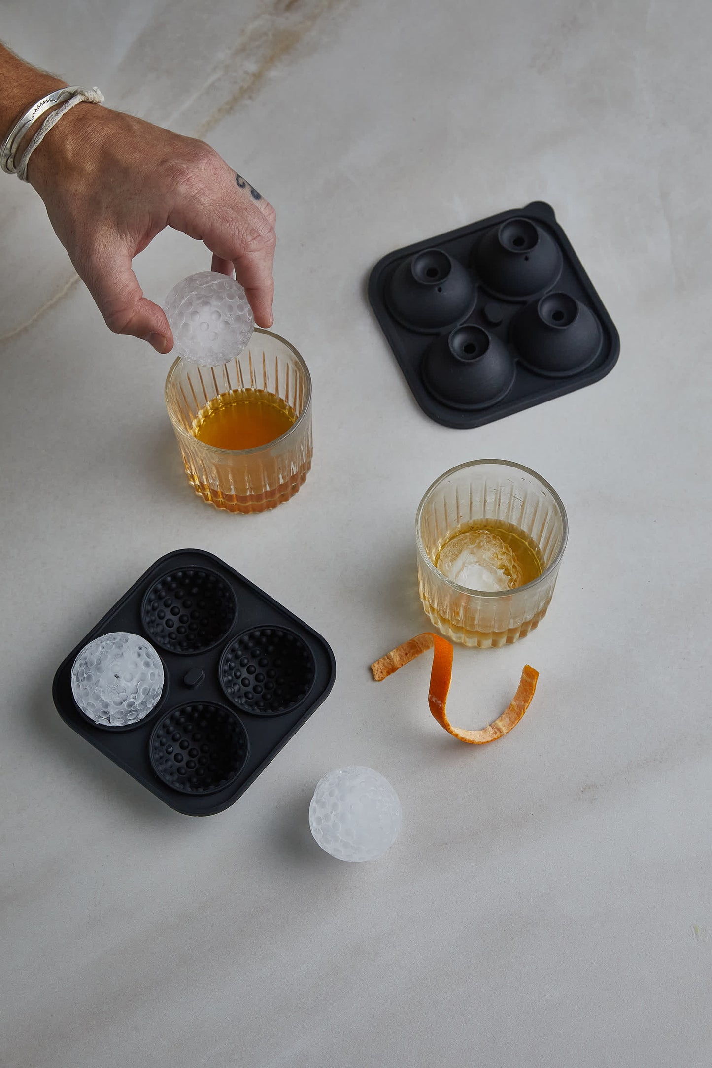 A hand drops a golf ball-shaped ice cube, made with the Golf Ball Ice Mould, into whiskey—beside silicone moulds, an extra glass, and an orange twist—the ideal touch for any golf enthusiast.