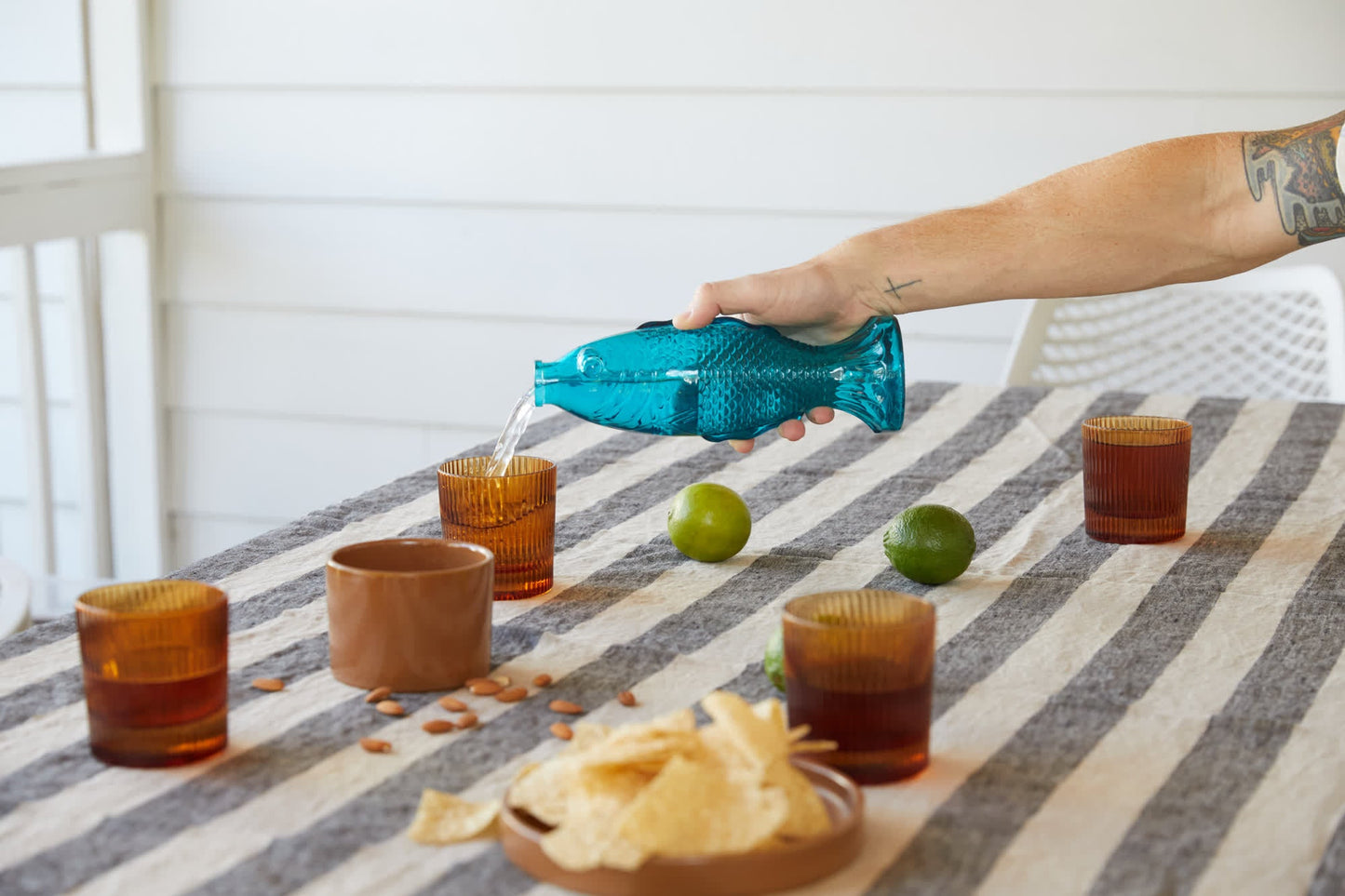 A person pours water from the Fish Decanter into a cup on a striped table decorated with snacks, limes, and three amber glasses.