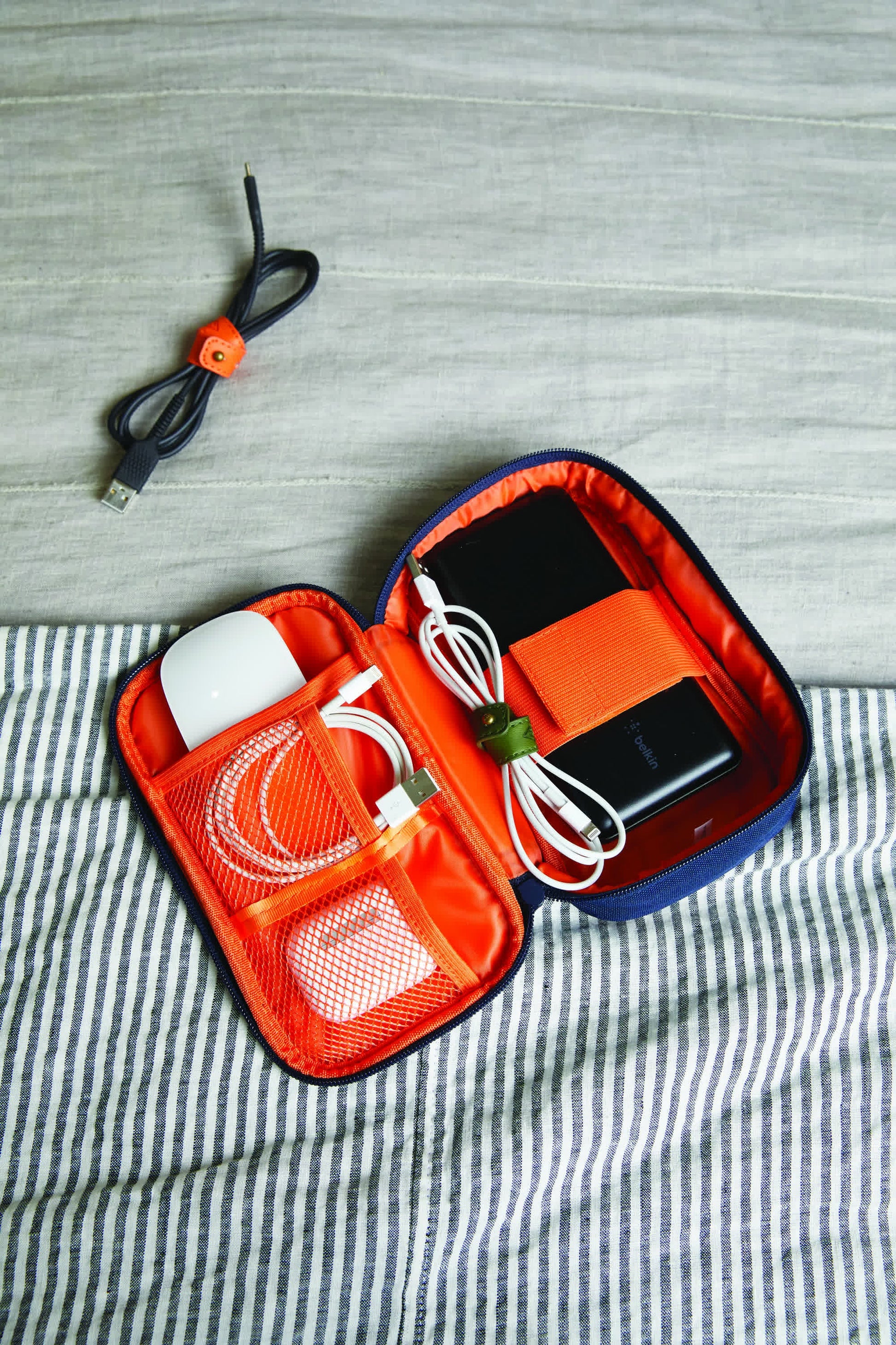 The Tech Tidy Case, featuring an orange lining, securely holds electronic accessories like a white mouse, cables, and devices. A nearby cable on a striped surface shows its ability to keep workspaces organized and clutter-free.