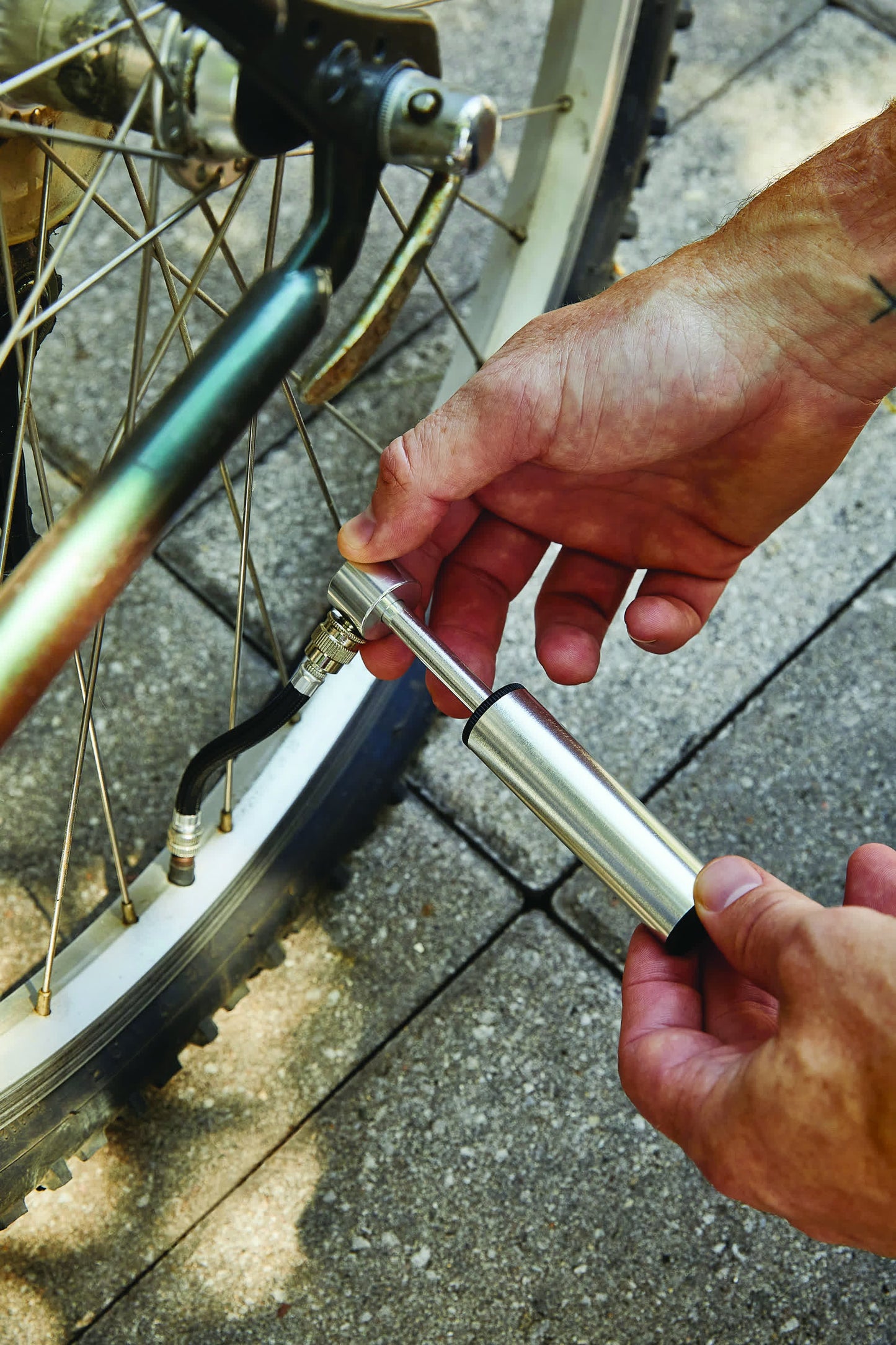 A person uses a Mini Bike Pump with valve adapters to inflate a bicycle tire on a paved surface.