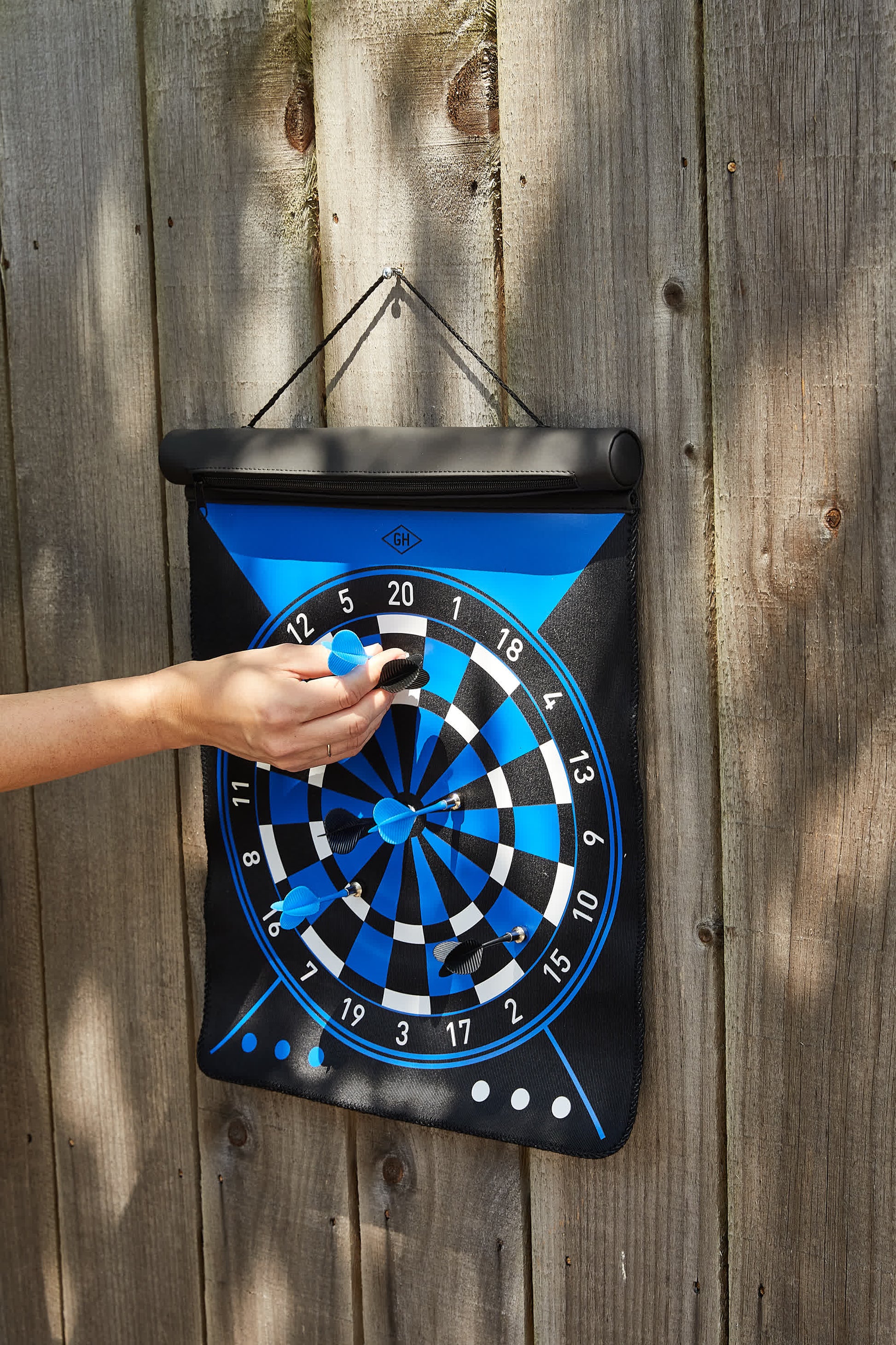 A person’s hand places a blue dart on the Dartboard Roll magnetic dartboard, which hangs on a wooden fence. Four blue darts are already on the board, showcasing fun and skill with Dartboard Roll.