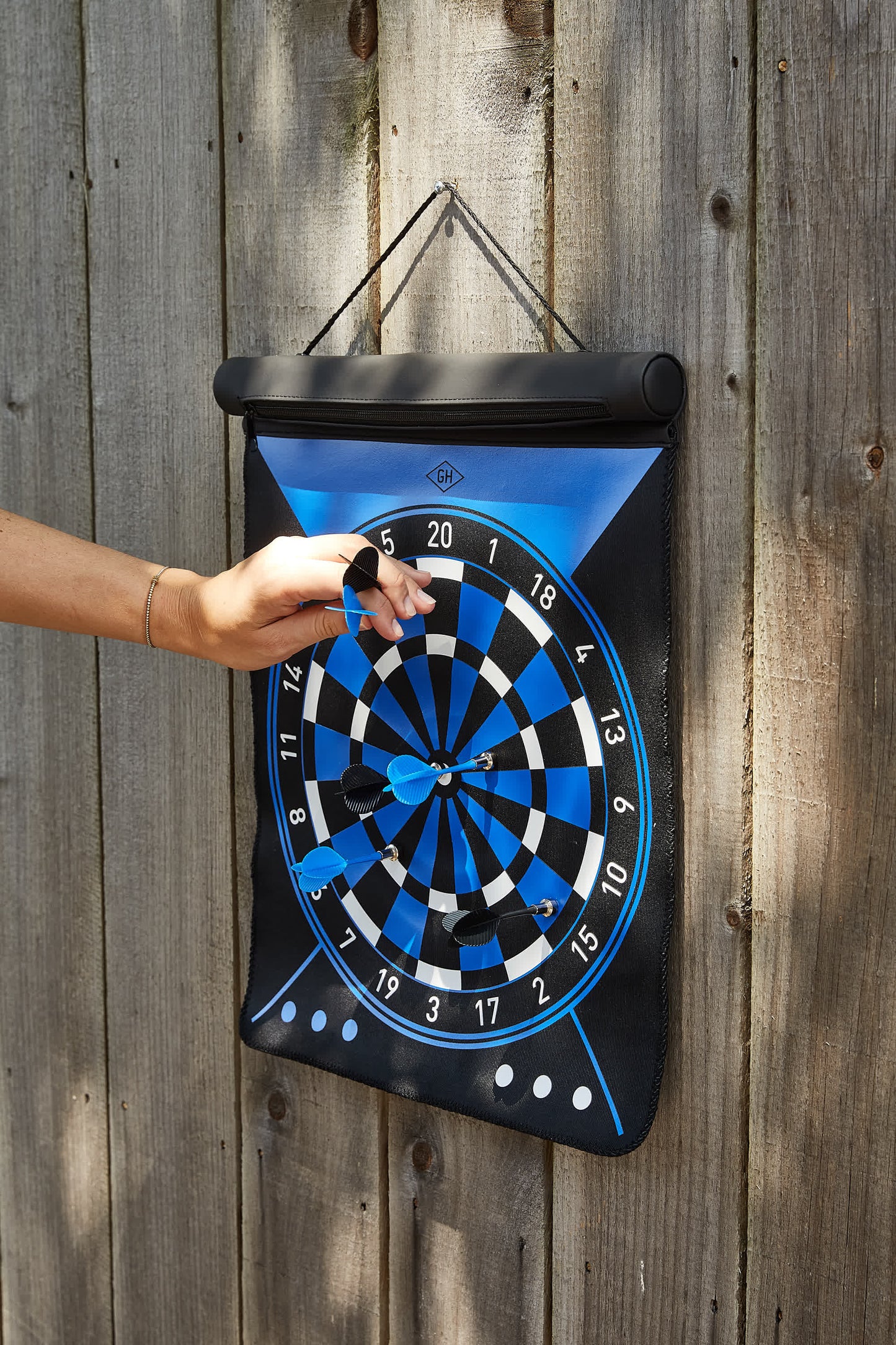 A hand removes a dart from the blue and black Dartboard Roll hanging on a wooden fence, with several darts stuck in it—demonstrating impressive hand-eye coordination.