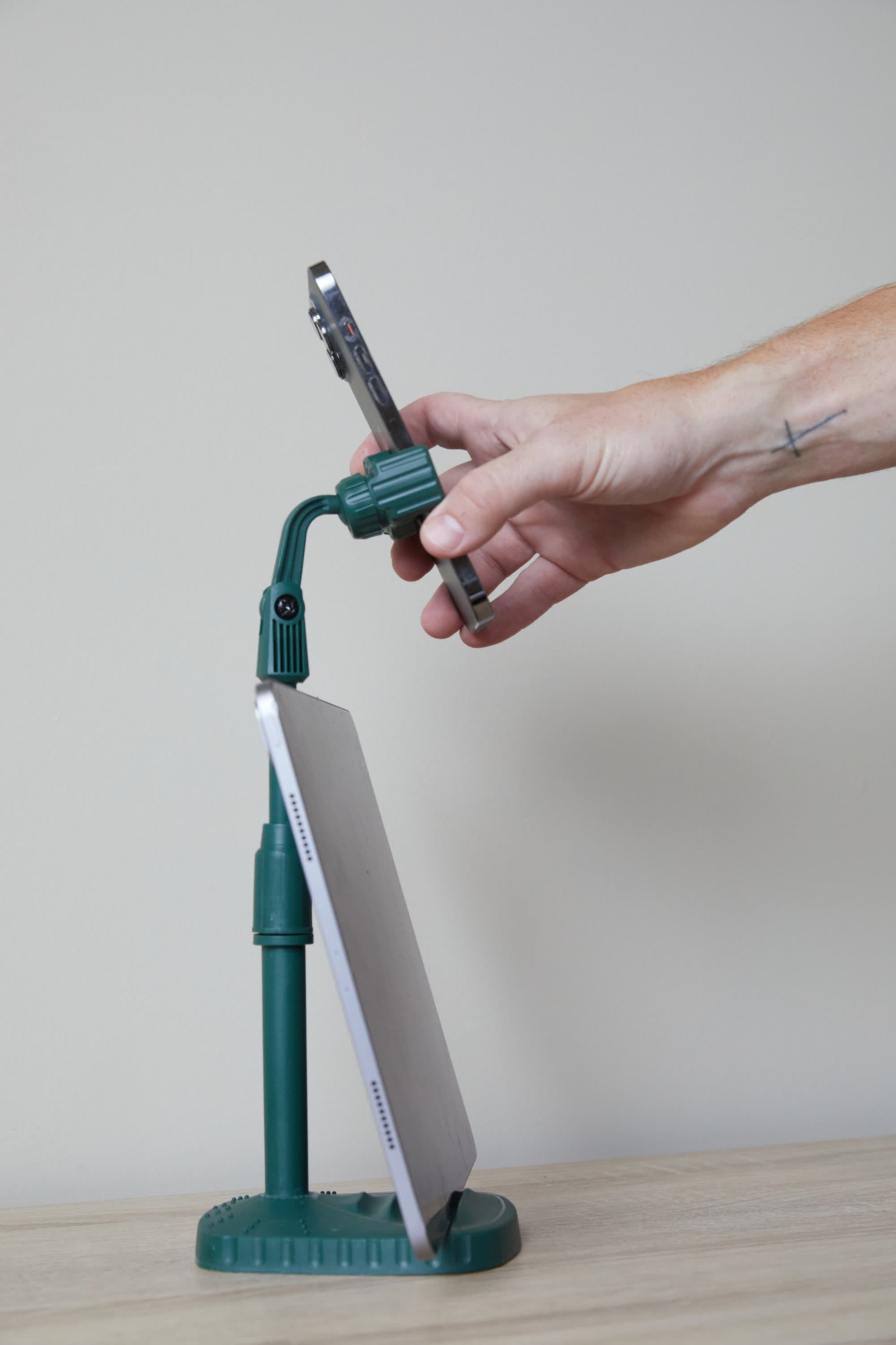 A hand adjusts a smartphone mounted above a tablet on the Phone Tripod—a versatile green desk accessory—against a plain light background.