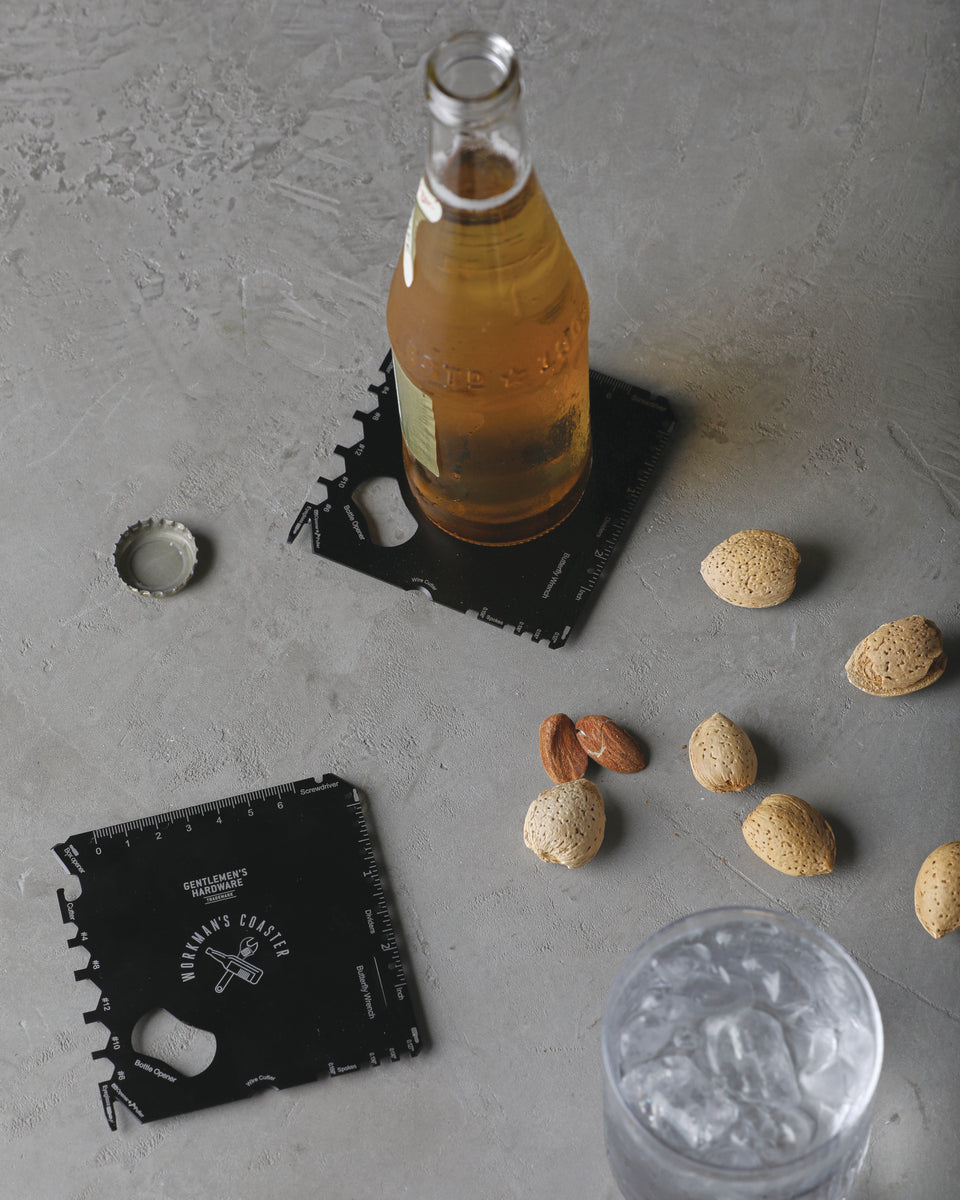 A bottle of beer and loose almonds rest on a gray surface. A glass of ice water, a bottle cap, and two Workmans Coaster sets are visible, with the coasters being used under the drinks.