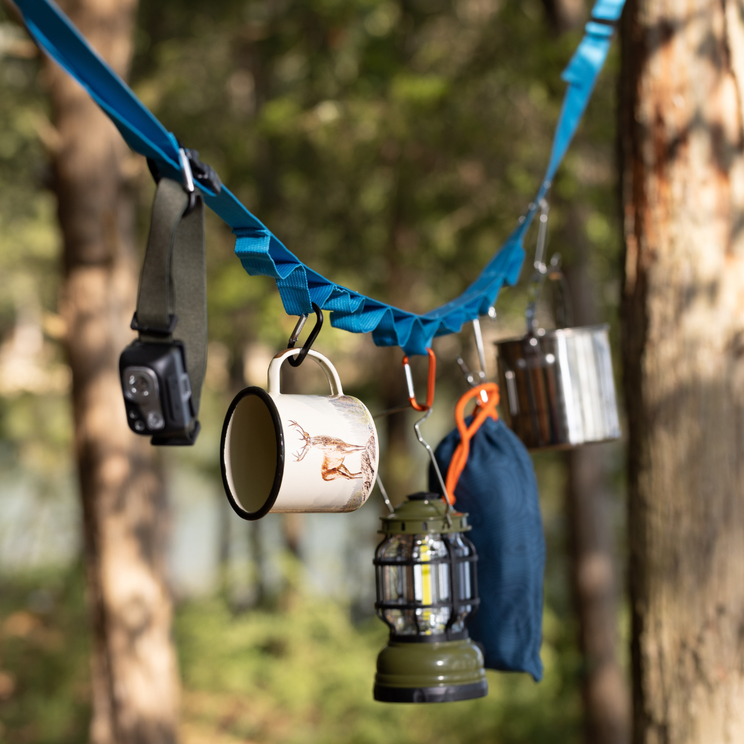 The Hanging Tent Tidy hangs between trees in a forest, keeping your gear organized by holding items like a headlamp, mug, lantern, metal container, and blue pouch.