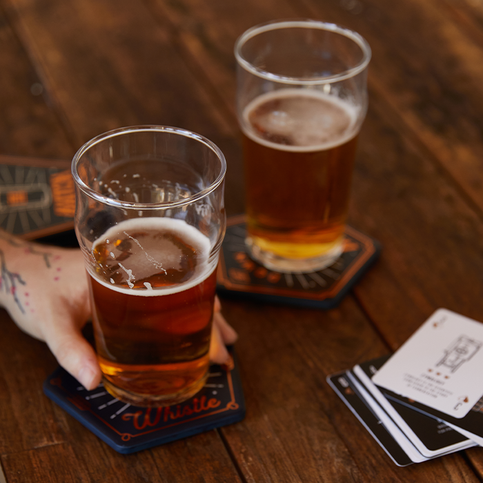 Two glasses of beer rest on the Beer Coasters, Set of 4 from Gentlemen’s Hardware atop a wooden table with playing cards; a tattooed hand holds one glass.