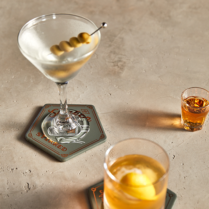 A martini with olives, an old fashioned with a lemon twist, and a shot of whiskey sit on the Cocktail Coasters, Set of 4, adding elegance to your barware collection on a light stone surface.
