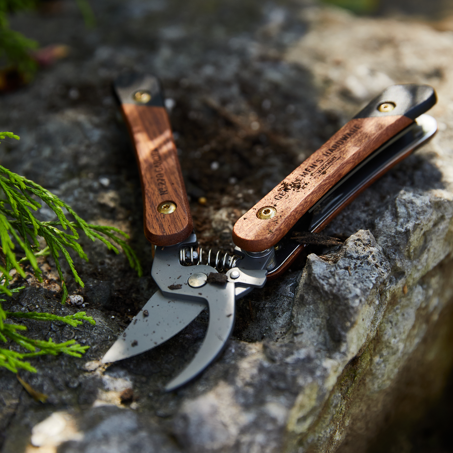A Garden Multi Tool with wooden handles lies open on a stone surface near green foliage.