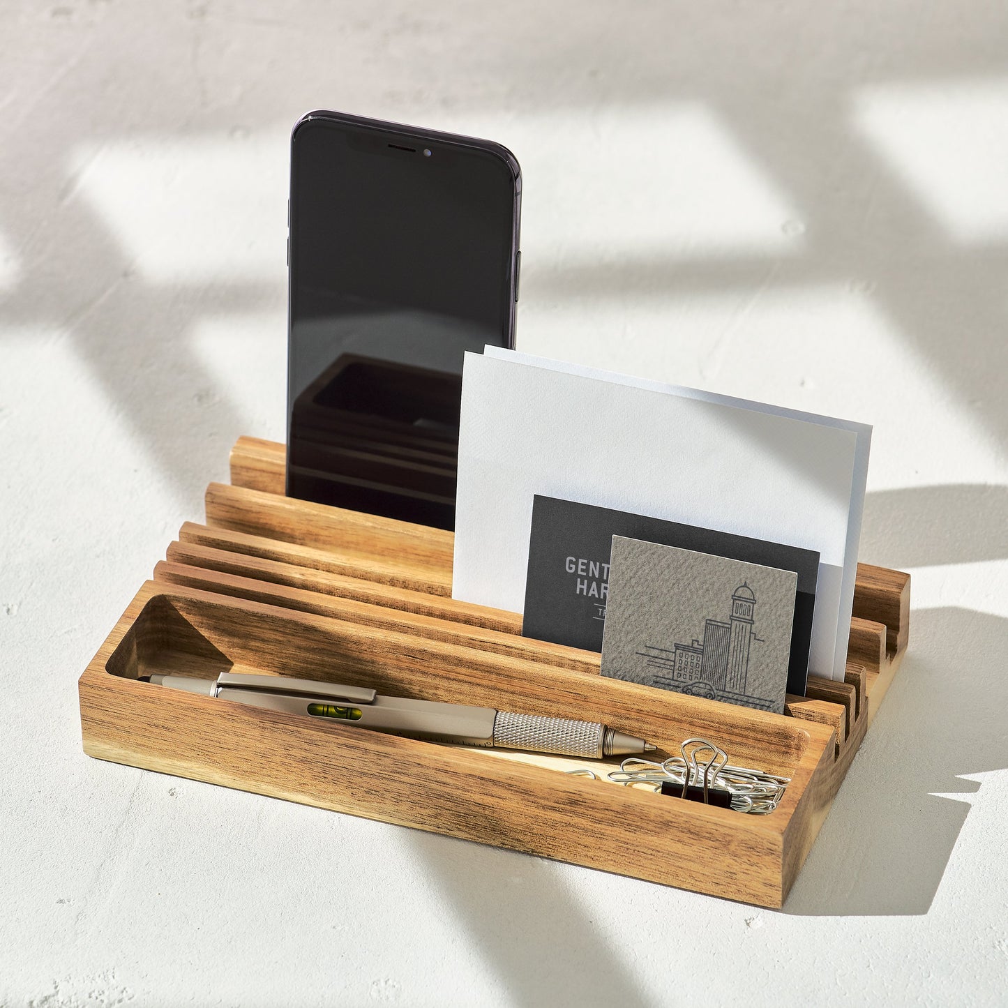 The Wooden Desk Organizer & Phone Stand neatly holds a smartphone, envelopes, card, pen, and paper clips, arranged on a white surface in natural light.