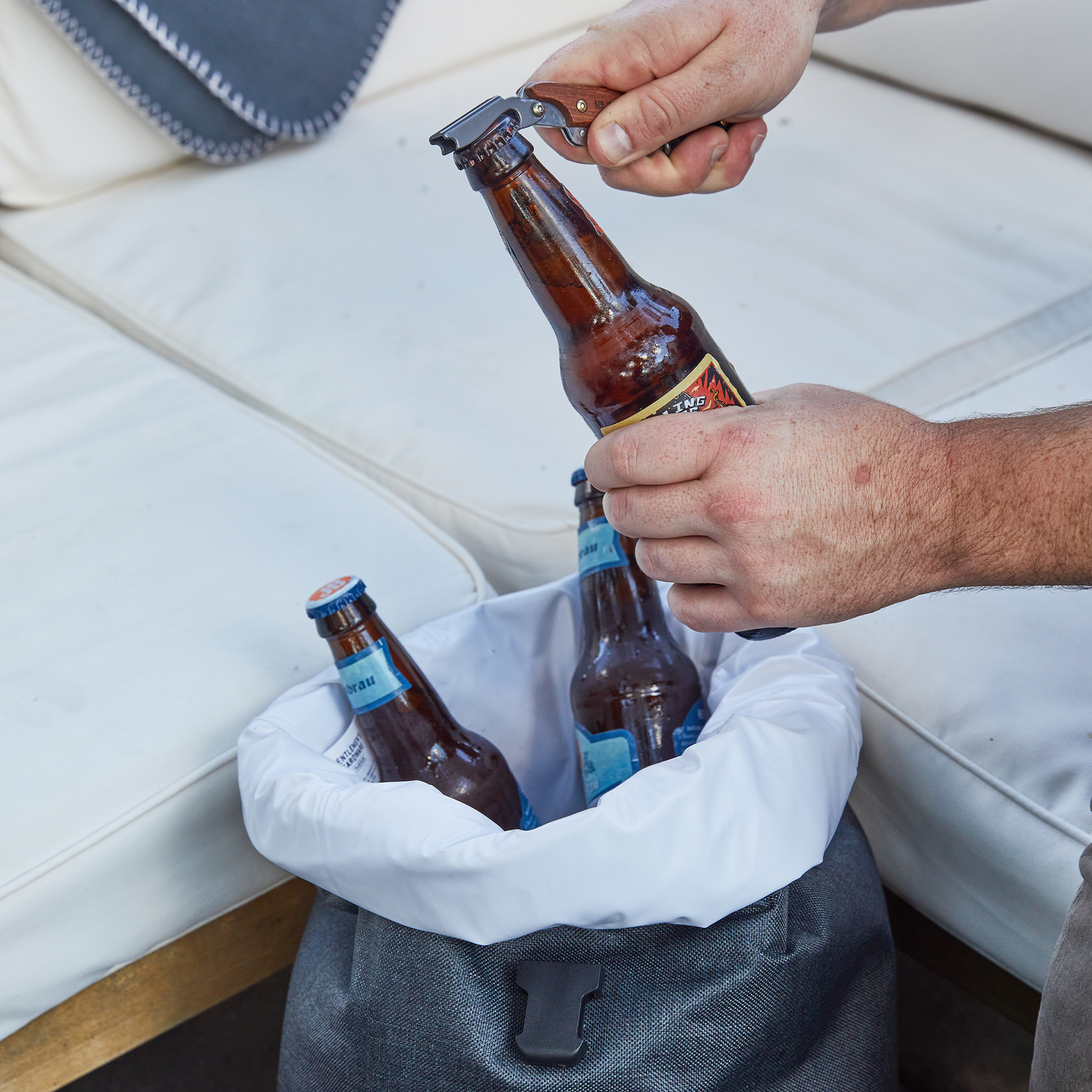 A person uses a bottle opener to open a beer, while two more bottles rest in an Insulated Cooler Backpack placed on a white outdoor couch.