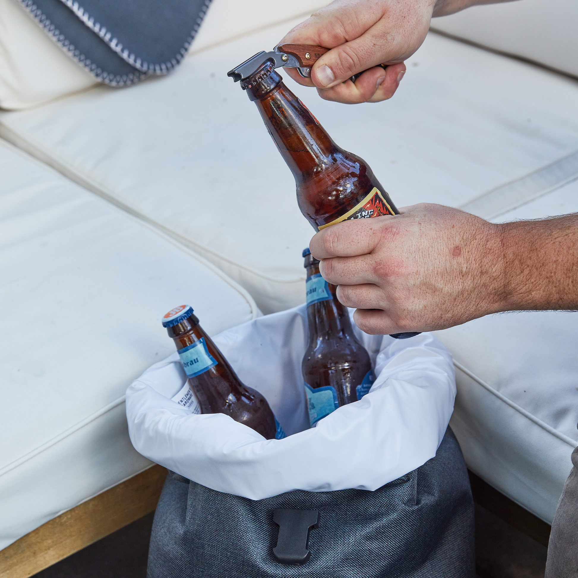 A person uses a bottle opener to open a beer, while two more bottles rest in an Insulated Cooler Backpack placed on a white outdoor couch.