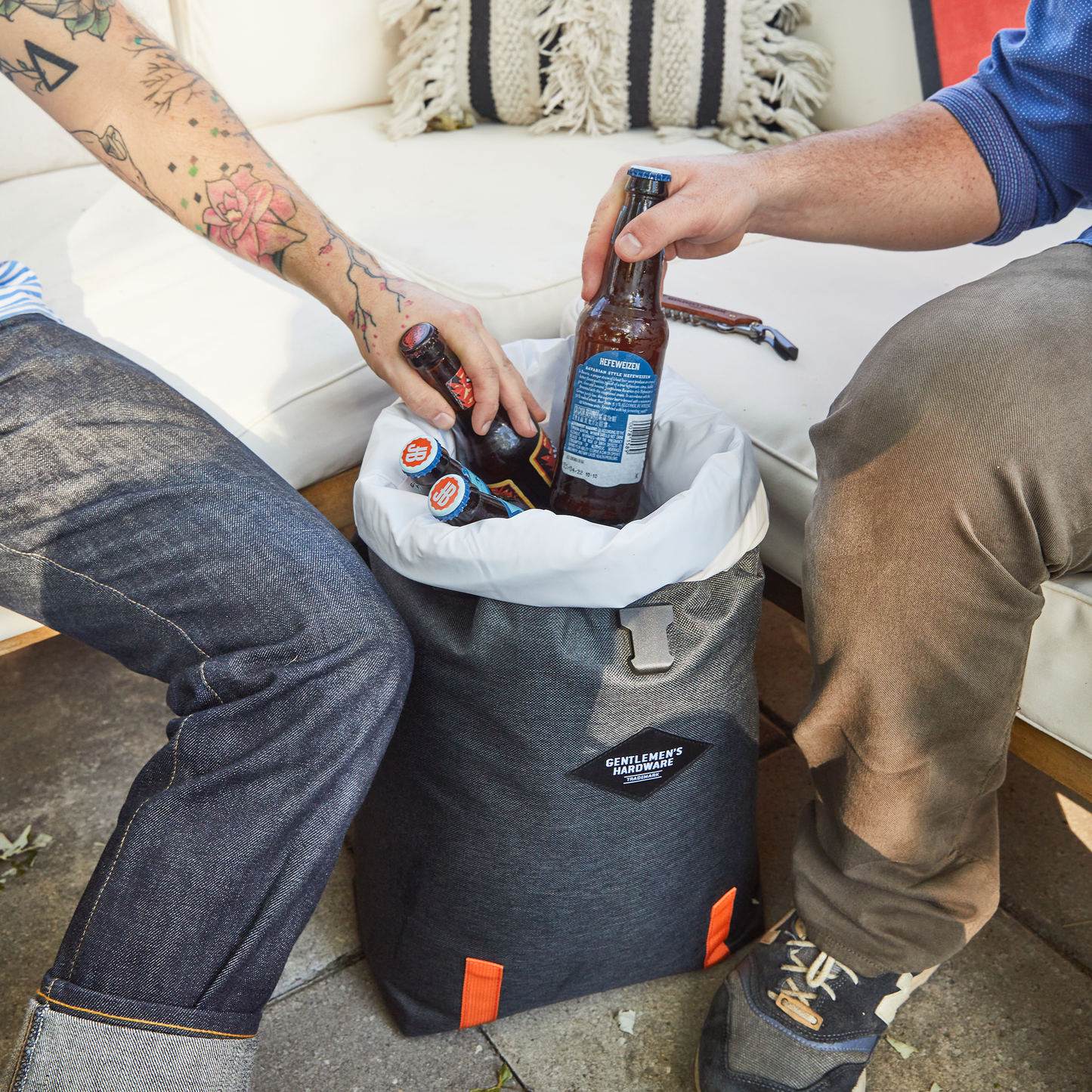 Two people sit on a couch, placing bottles of beer into an Insulated Cooler Backpack featuring a gray exterior, white liner, and orange accents.