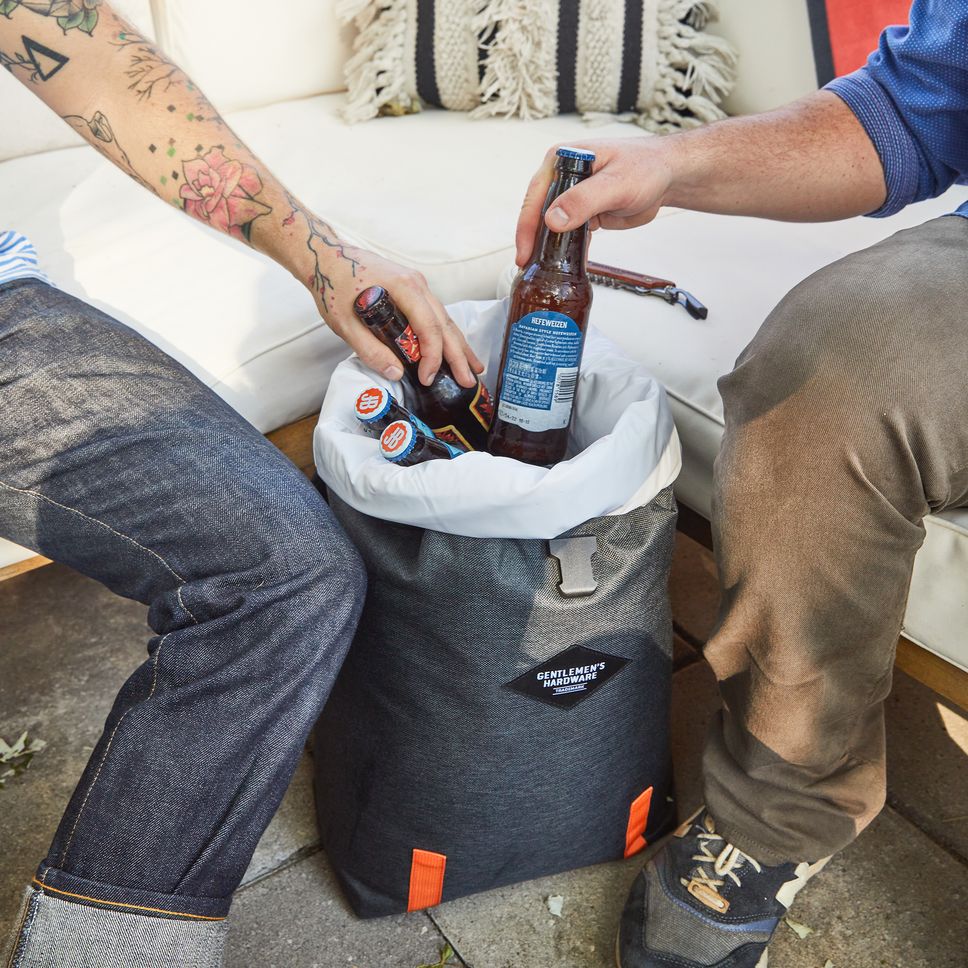 Two people sit on a couch, placing bottles of beer into an Insulated Cooler Backpack featuring a gray exterior, white liner, and orange accents.