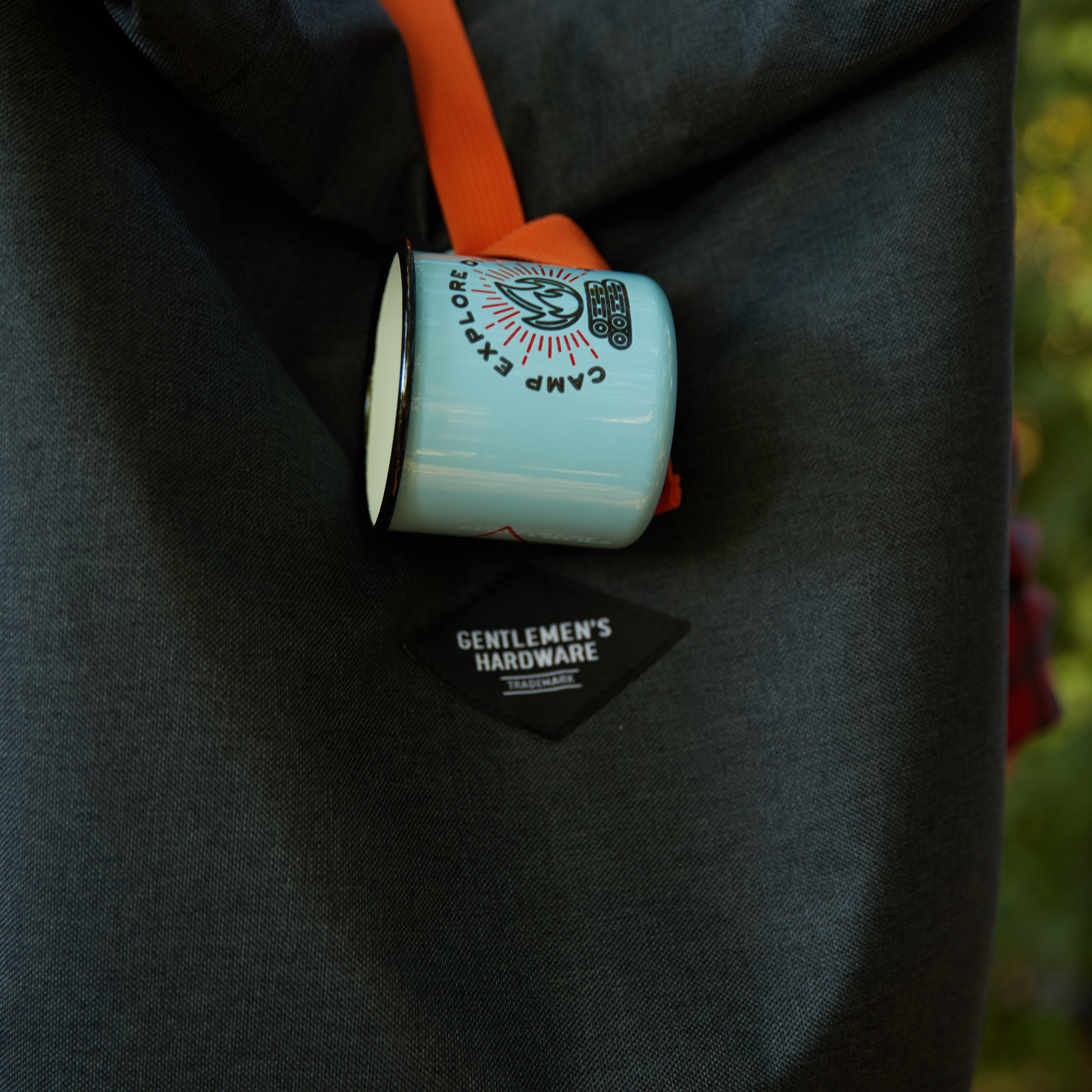 An Enamel Mug - Camp. Explore. Discover. 11 fl oz with a camp-themed design hangs from a dark gray Gentlemens Hardware backpack, secured by an orange strap.