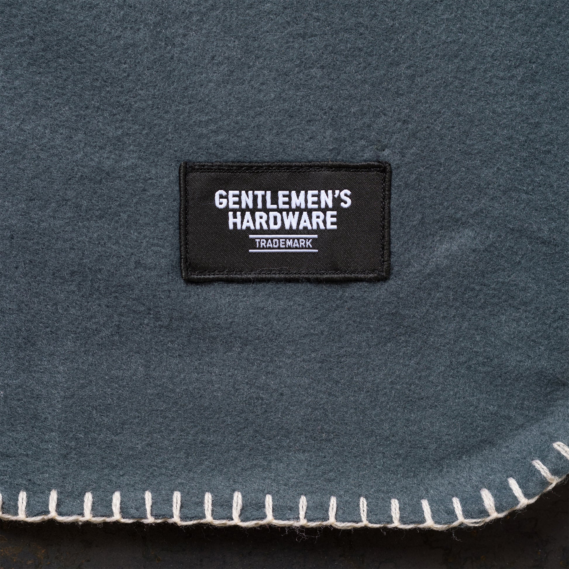The Camping Blanket is a lightweight, dark grey fabric with stitched edges and a rectangular black patch labeled Gentlemen’s Hardware Trademark in white—ideal for outdoor adventures.