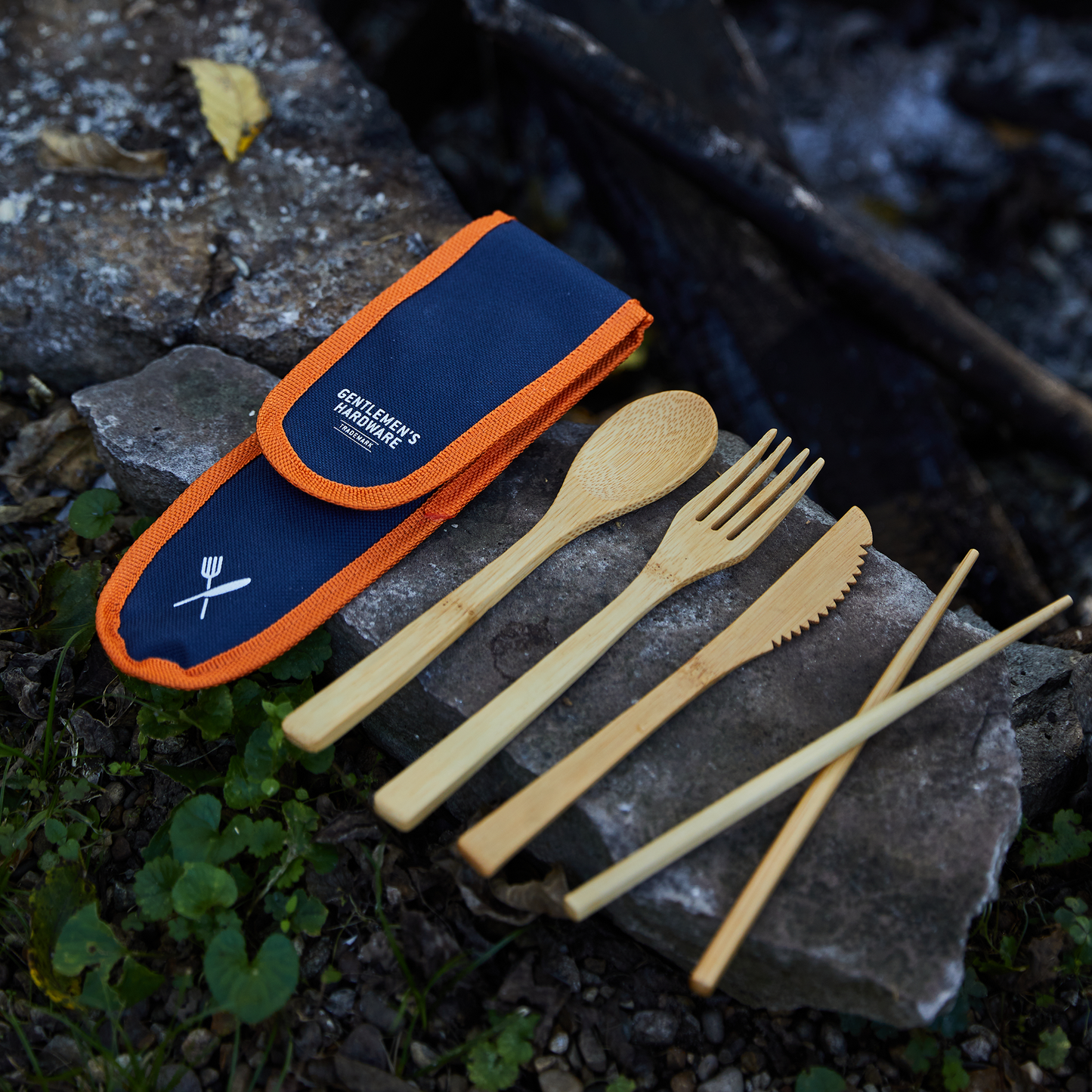 The Travel Bamboo Cutlery Set includes reusable chopsticks, knife, fork, spoon, straw with cleaning brush, all shown on rocks next to a navy and orange carrying case.