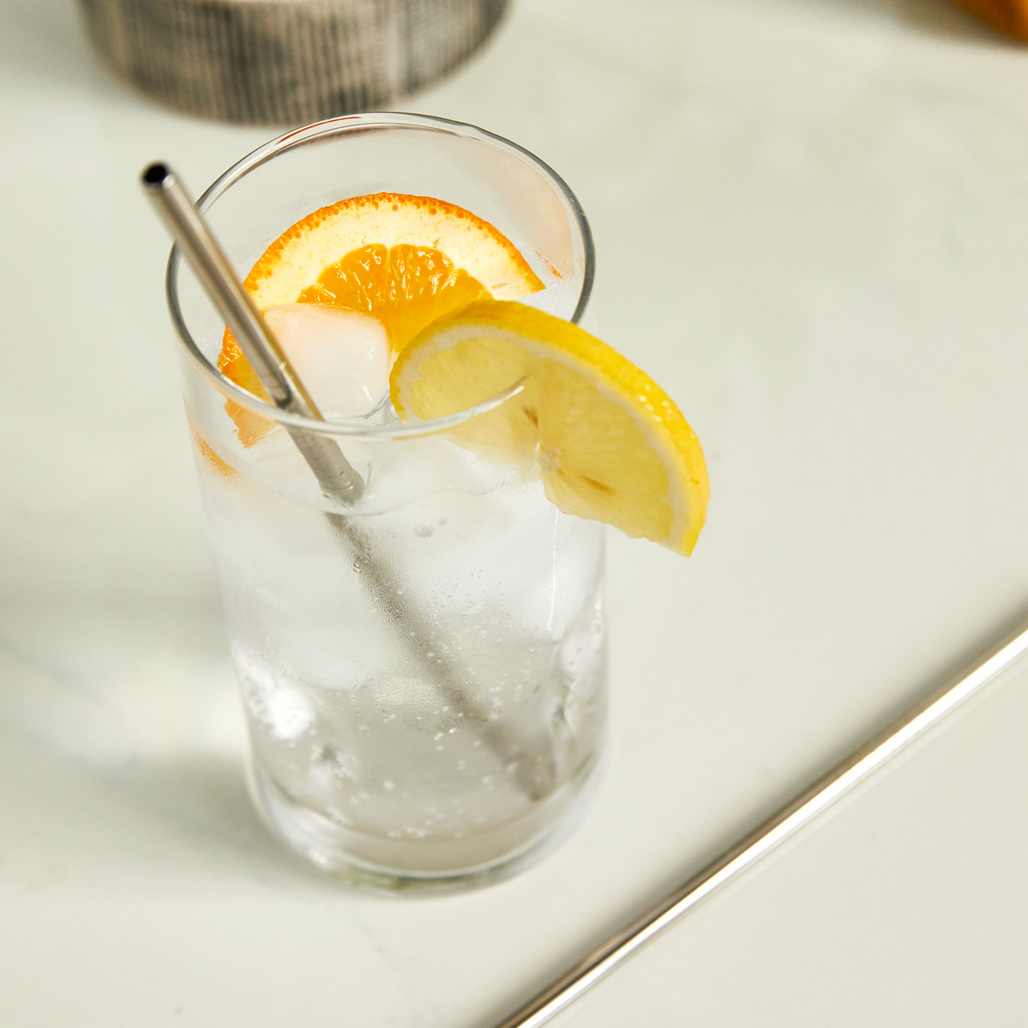 A glass of iced water with lemon and orange slices sits on a light surface, featuring a portable straw from the Travel Metal Straw Set.