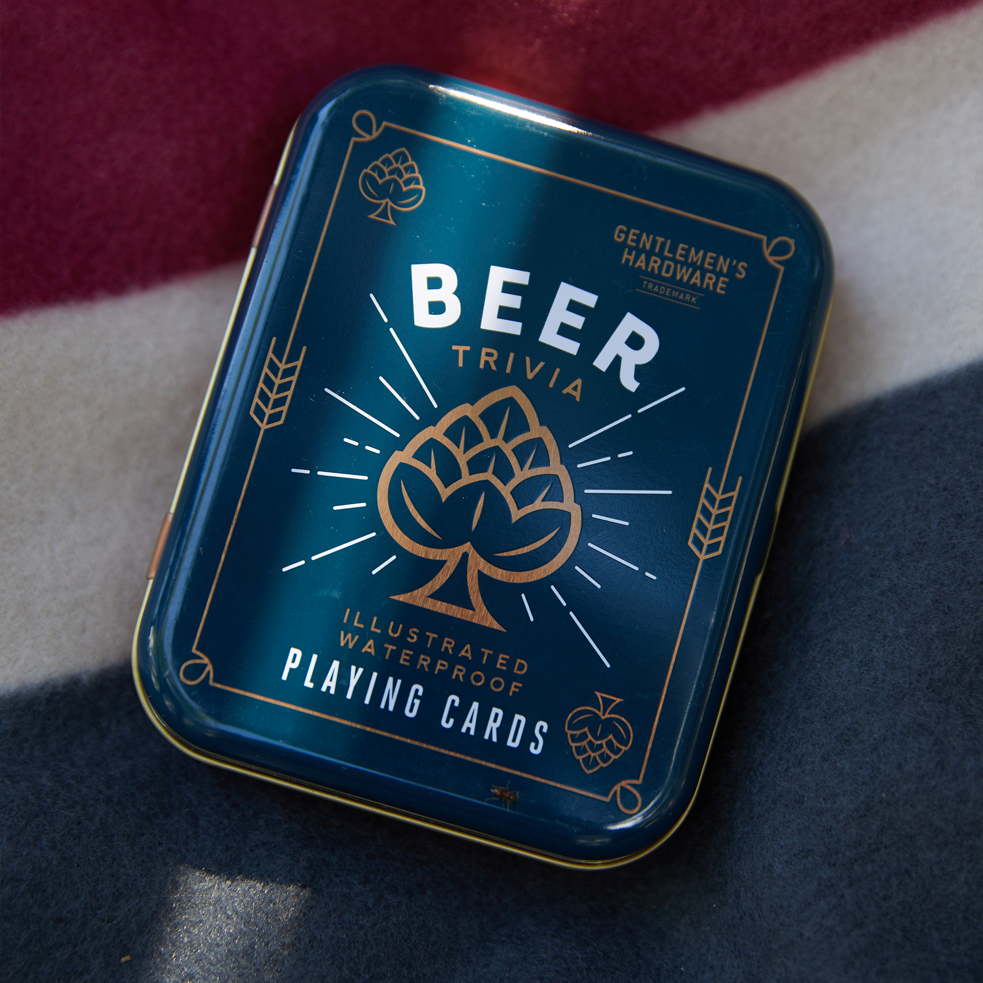 A metal tin labeled Beer Playing Cards sits on striped fabric, containing waterproof cards perfect for trivia fans.