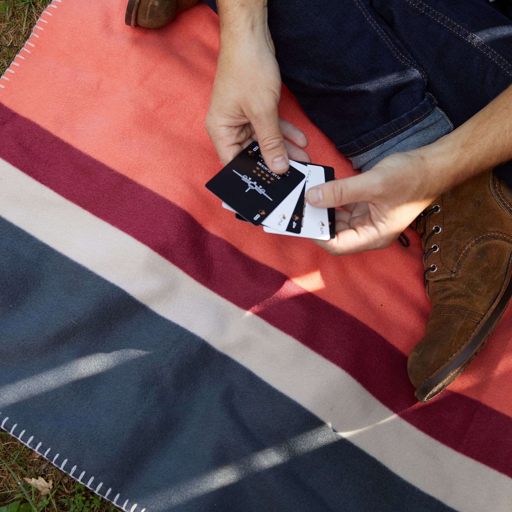 A person sits outdoors on a striped blanket, holding Beer Playing Cards in their hands—only hands, part of legs, and a brown boot are visible. Its the perfect setup for cards from the metal tin travel case.