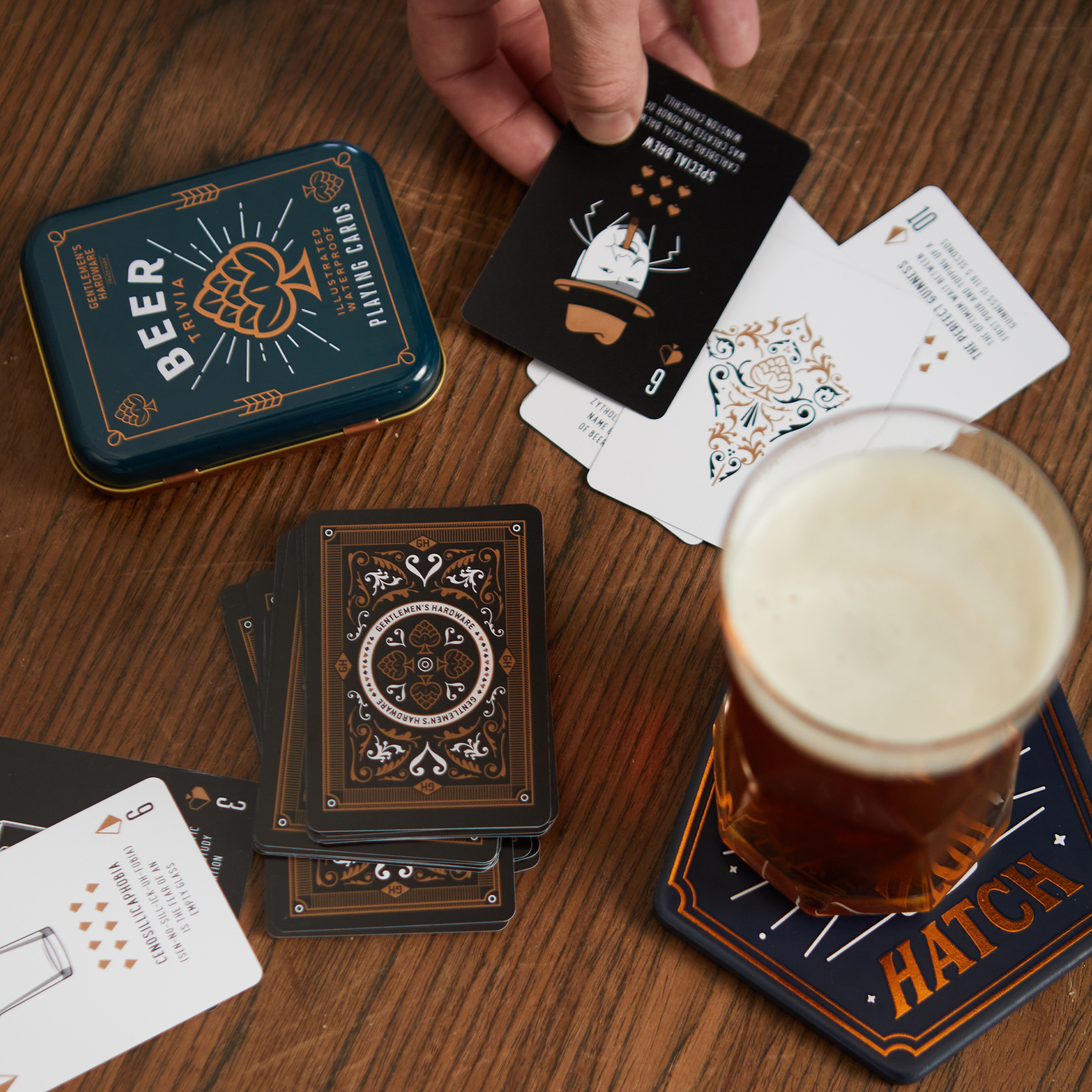 A hand holds a Beer Playing Card above a table featuring the Beer Playing Cards’ metal tin travel case, waterproof cards dealt out, and a glass of beer on a HATCH coaster.