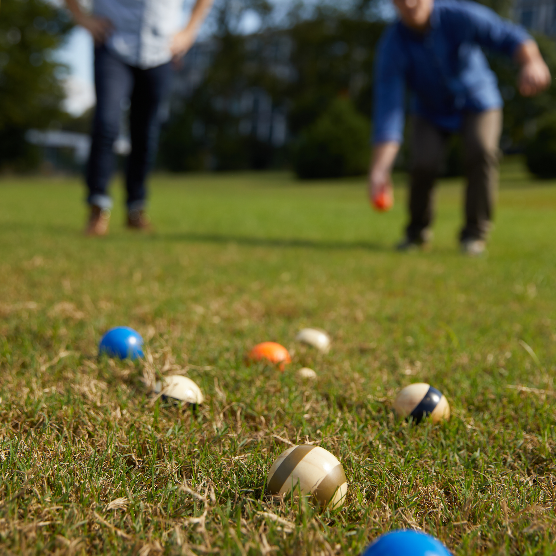 Two people enjoy a game of bocce on the lawn using the Bocce Ball Set with Travel Case; several colored and striped balls are scattered nearby, making this classic yard game ideal for beginners.
