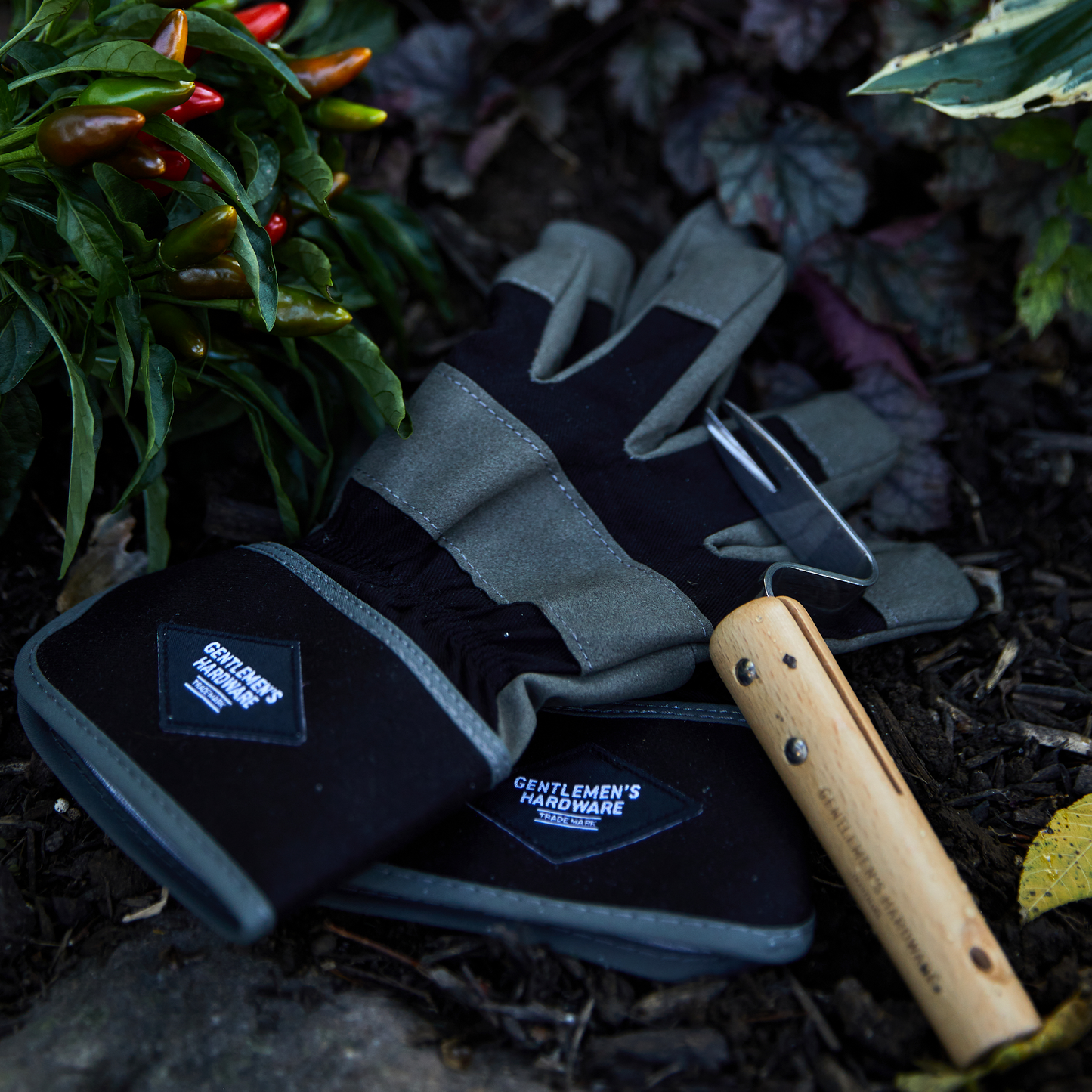 The Garden Gloves and Root Lifter, essential gardening tools, rest on soil near green and red chili peppers and leafy plants.