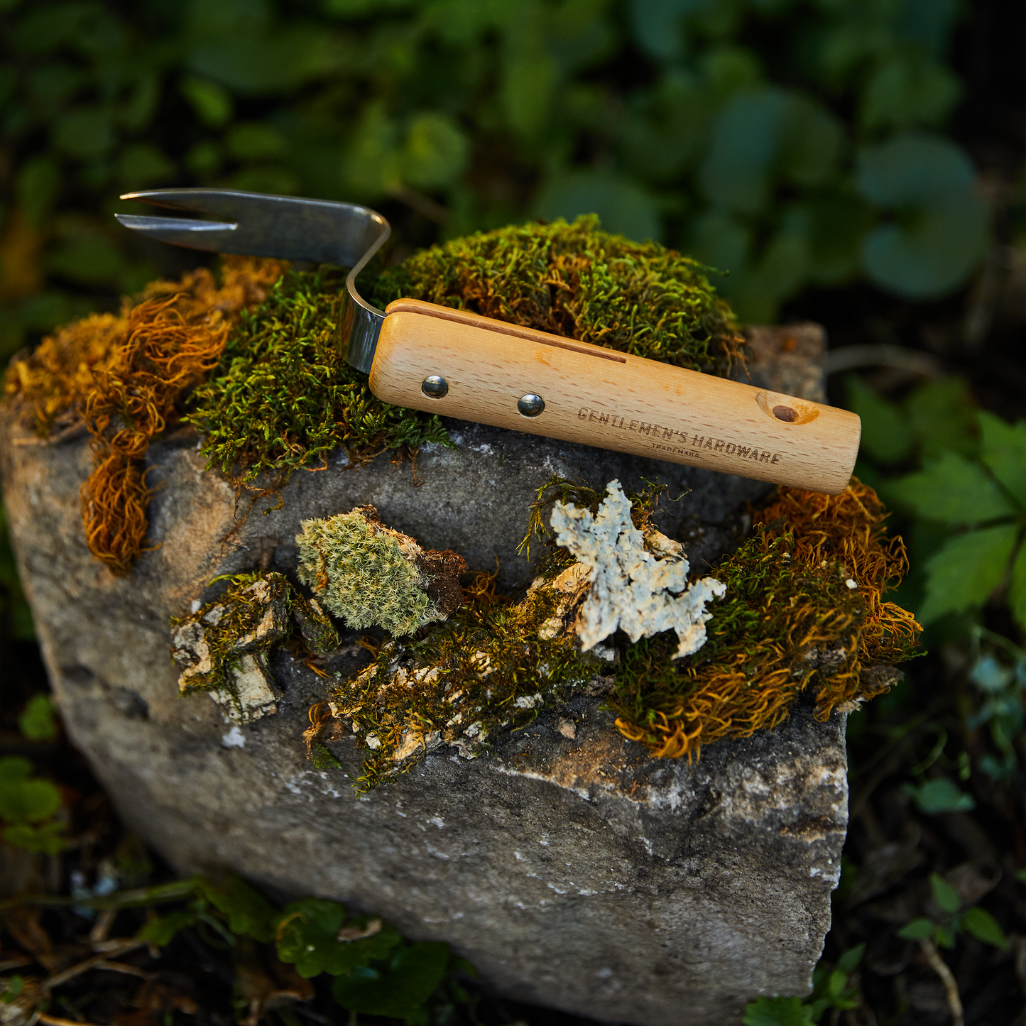 A small garden weeder with a wooden handle labeled Gentlemens Hardware rests on a mossy stone outdoors, the perfect companion to Garden Gloves and Root Lifter.