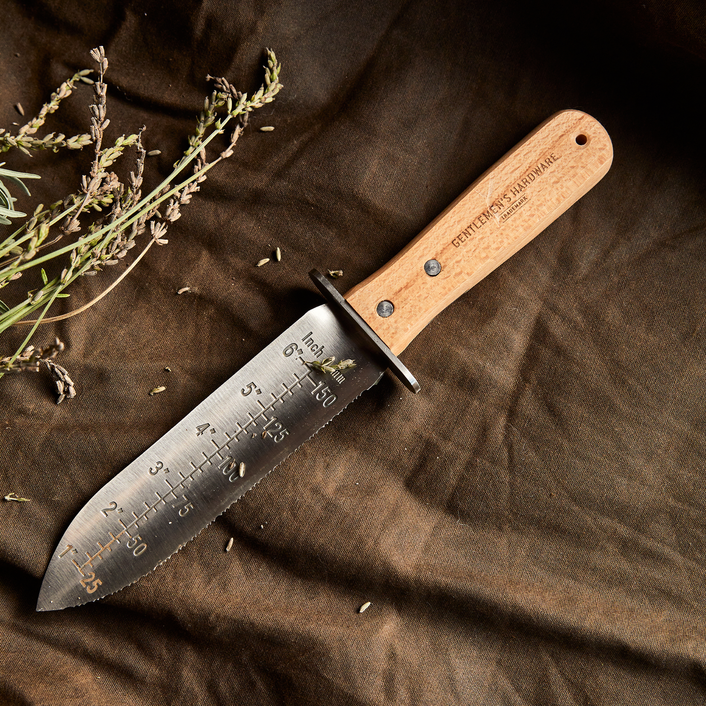 The Hori Hori Knife, featuring a wooden handle and measurement markings on its stainless steel blade, rests on brown fabric beside dried herbs.