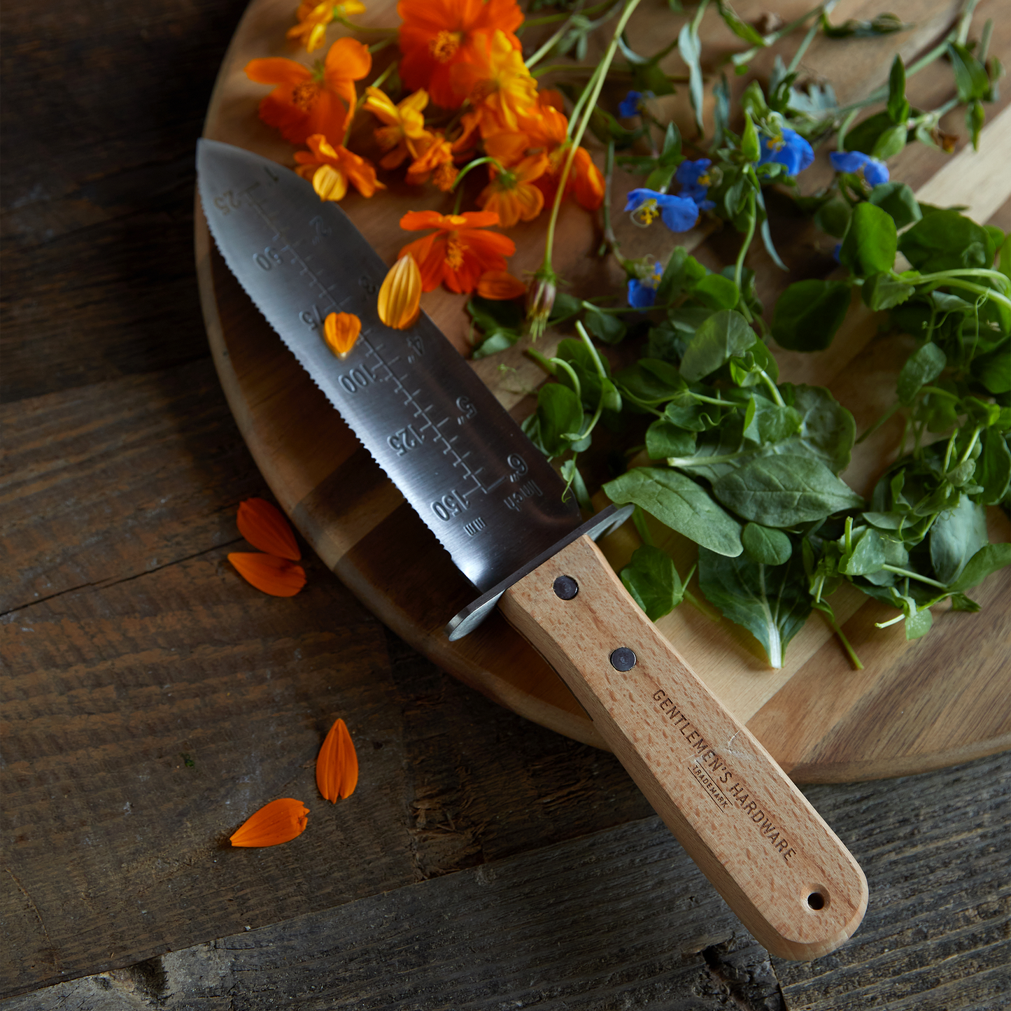 A Hori Hori Knife with a stainless steel blade and measurement markings lies on a wooden board beside fresh greens and colorful edible flowers.