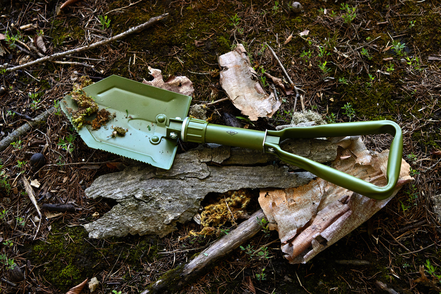 Olive green Folding Shovel resting on bark and moss