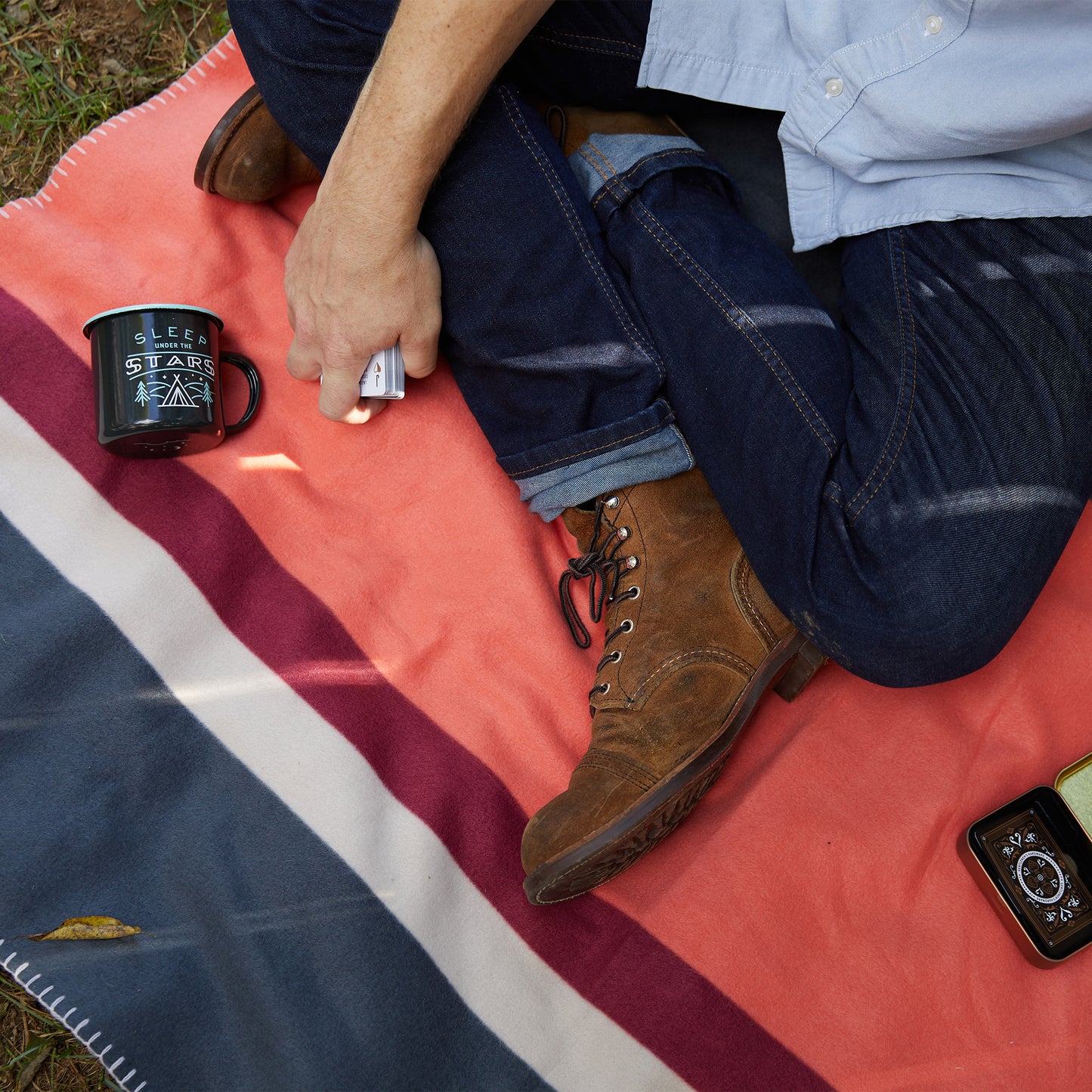 Relax outdoors on our Camping Blanket—lightweight and striped, its ideal for any adventure. Pair it with your favorite drink, just like the person sitting cross-legged in jeans and boots with a black mug by their side.