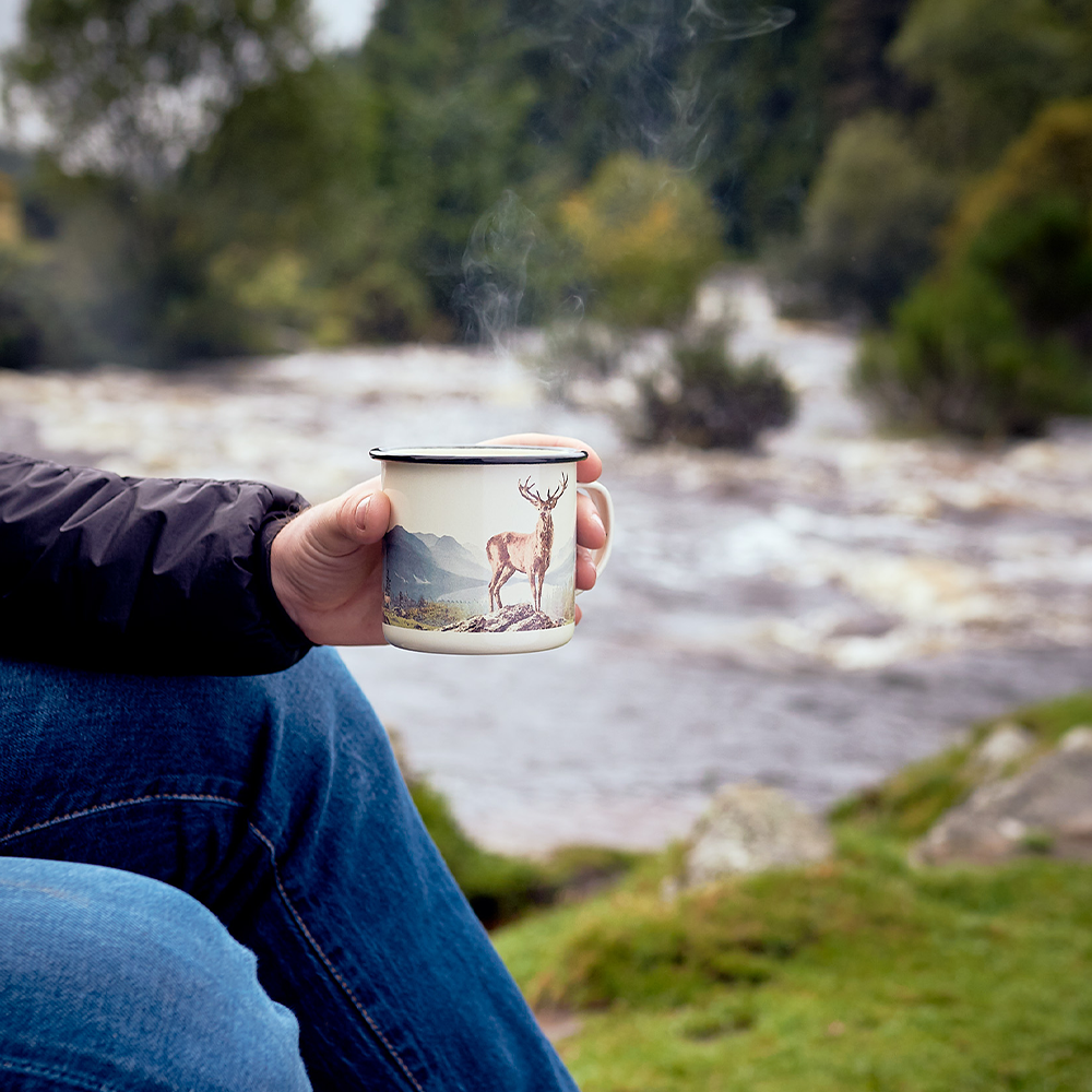 Someone sits by a river in nature, holding the Enamel Mug - Great Outdoors Deer 17 fl oz. Steam rises from the mug, reflecting peaceful moments and adventure in the outdoors.