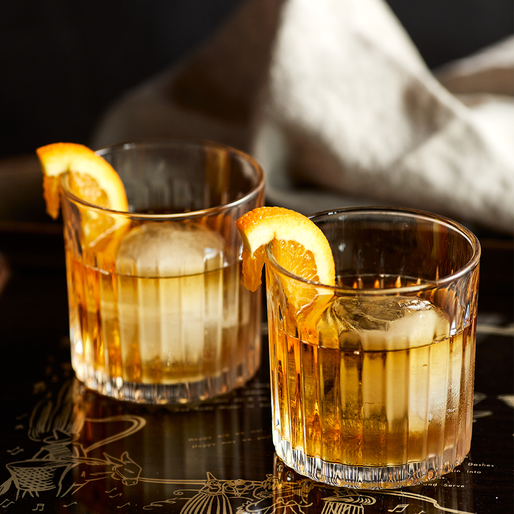 Two glasses of whiskey with large ice cubes and orange wedges on a dark surface—perfect inspiration to showcase your Whiskey Tumbler Glasses & Ice Stones Set.