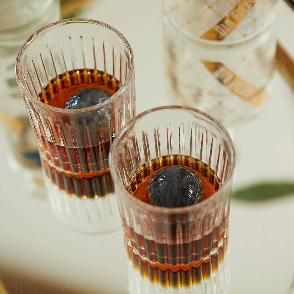 Two glasses from the Whiskey Tumbler Glasses & Ice Stones Set, filled with amber liquor and round ice spheres, rest on a reflective surface—a stylish choice for your home bar or as a thoughtful gift. A patterned bottle appears in the background.
